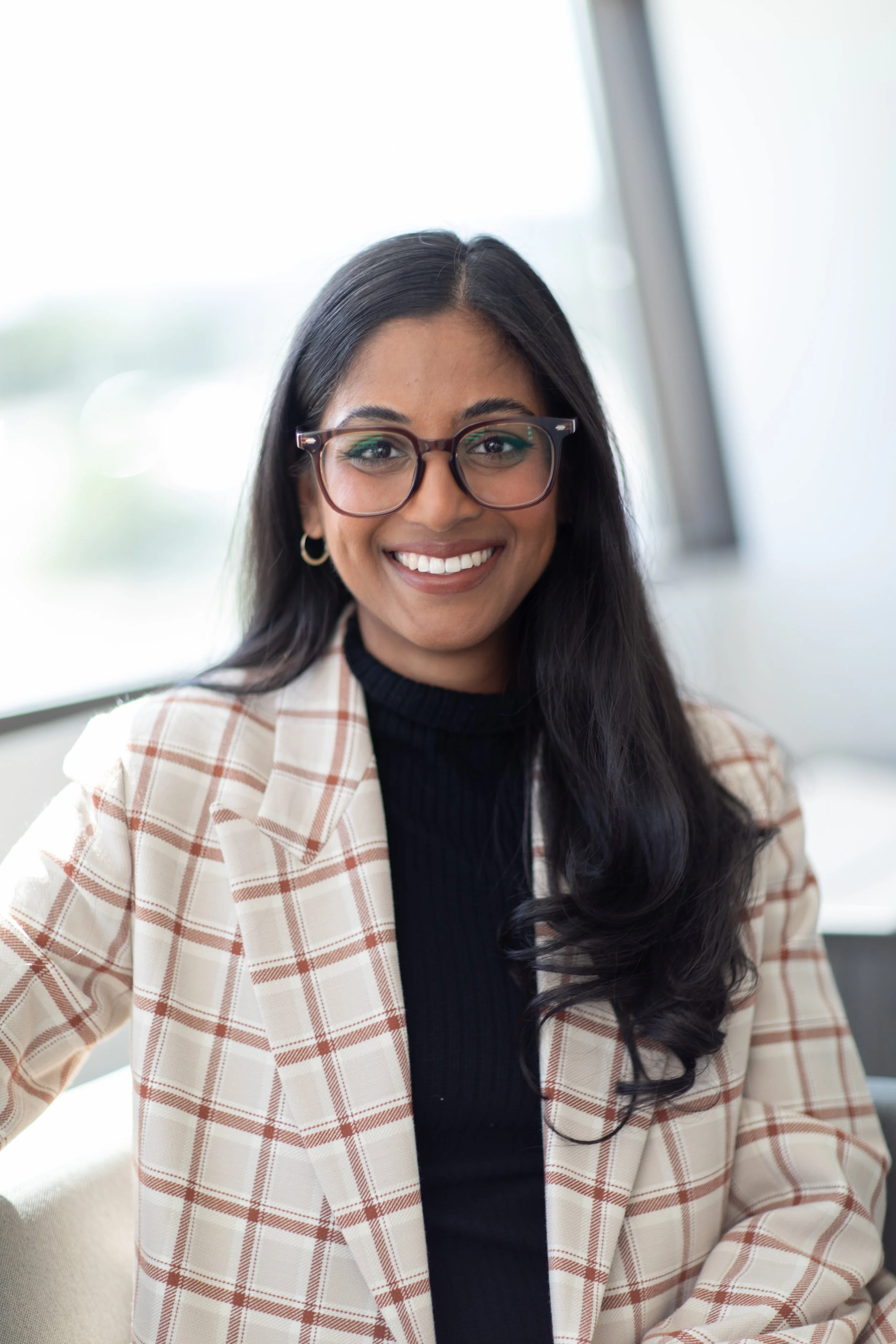 Smiling woman with long dark hair, glasses, hoop earrings, wearing a plaid blazer and black turtleneck, sitting near a window.