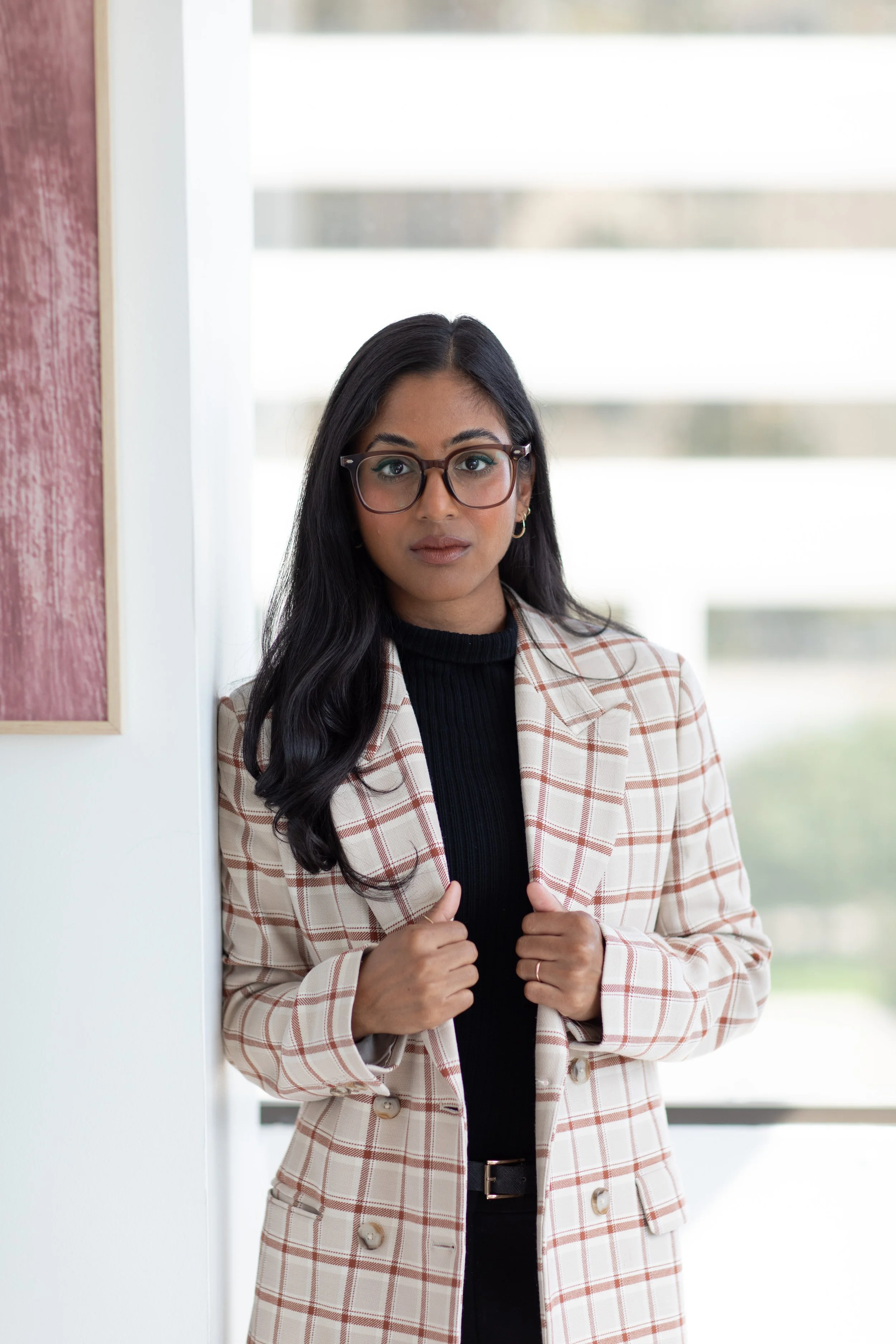 A young woman with dark hair, glasses, and light skin, wearing a plaid blazer and holding the lapels, standing indoors near a white wall with a blurred window background.