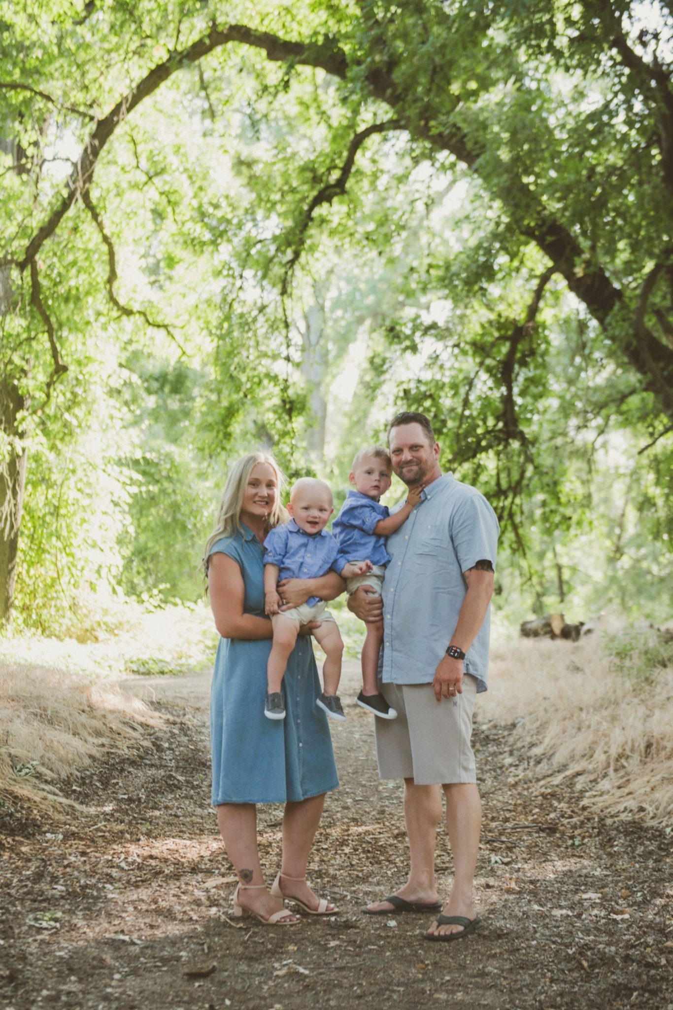 Family of four standing in a wooded outdoor area, smiling for a photo. The woman is wearing a blue dress and high heels, holding a young boy in a blue shirt and shorts. The man, dressed in a light blue shirt and shorts, is holding another young boy in a similar outfit. They are surrounded by tall trees with green leaves.