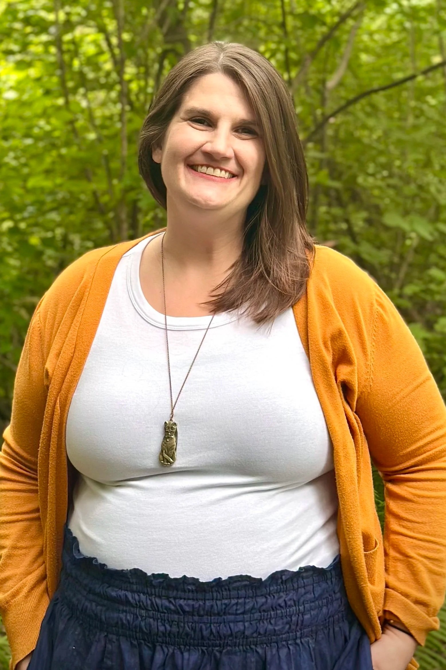 A woman with shoulder-length brown hair smiling outdoors among green trees, wearing a white shirt, an orange cardigan, a necklace with a cat pendant, and a dark skirt.