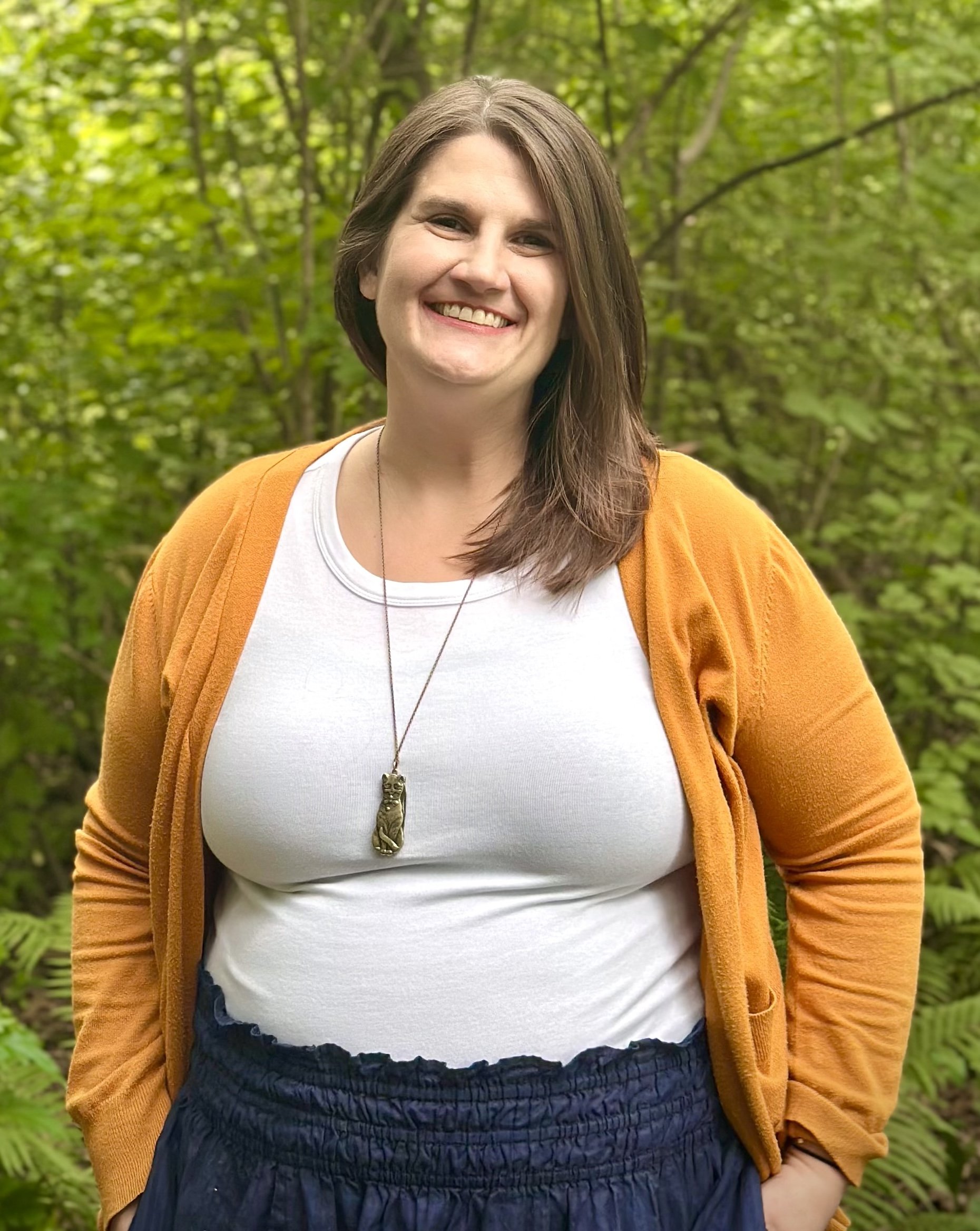 Meg Chuhran LICSW smiling in white shirt, mustard cardigan and navy skirt with trees behind