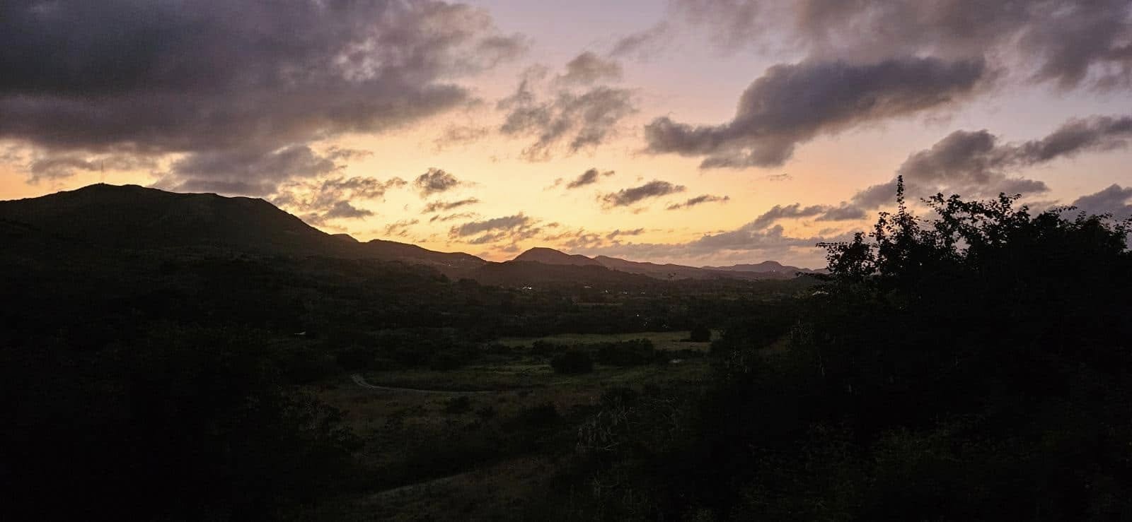 Silhouette of rolling hills and trees under a vibrant orange and purple sunset sky with scattered clouds.