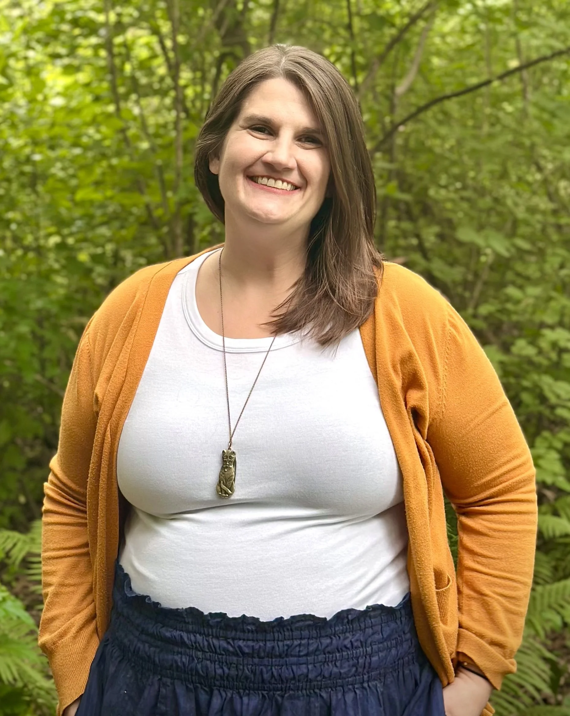 Meg Chuhran, LICSW, smiling with shoulder-length brown hair poses outdoors in front of a lush, green leafy background wearing a yellow cardigan, white shirt, and navy blue skirt. She is wearing a cat necklace.