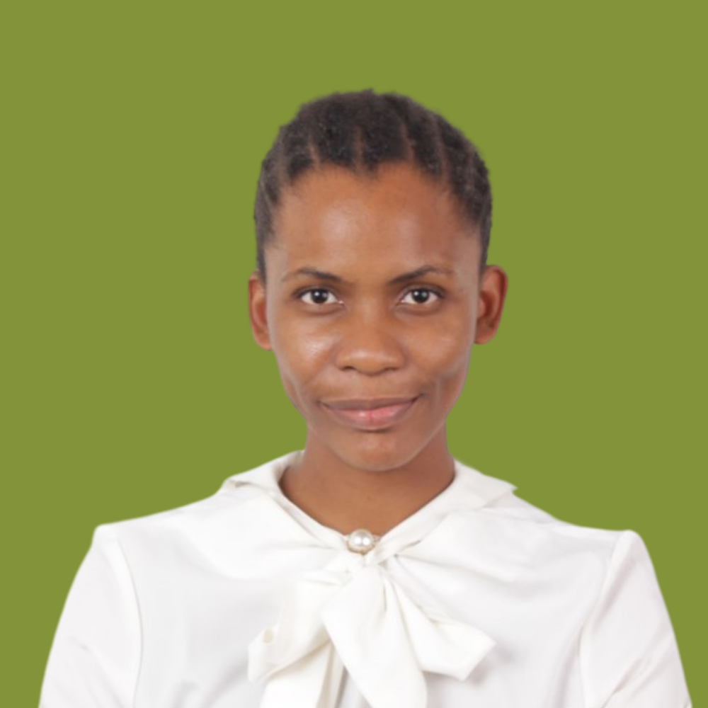 A woman with short, natural hair standing with her arms crossed, wearing a white blouse with a bow at the collar, against a plain green background.