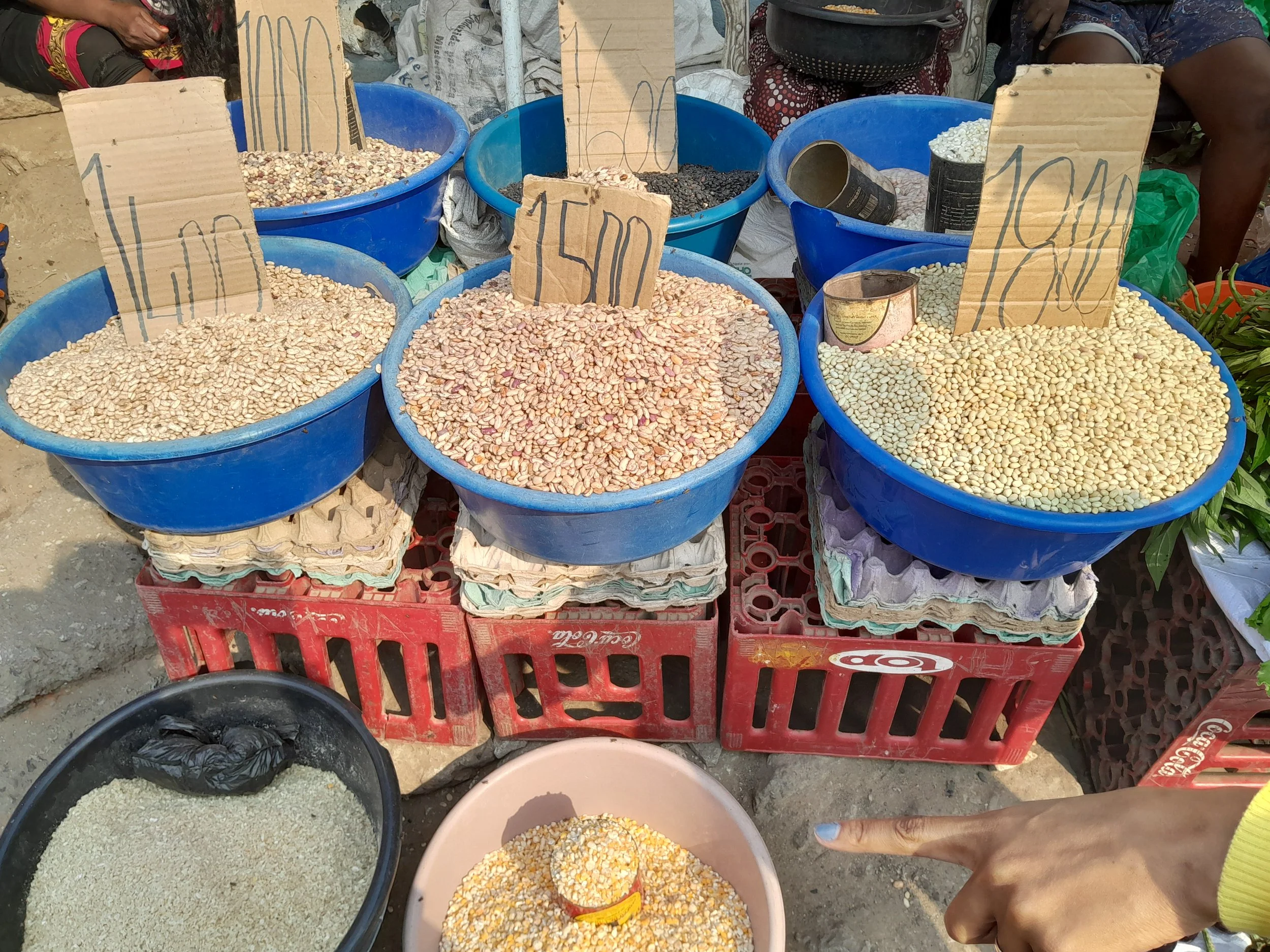 Multiple large blue buckets filled with various types of beans and grains at an outdoor market stall, some with handwritten cardboard signs indicating prices, with people and other market items in the background.
