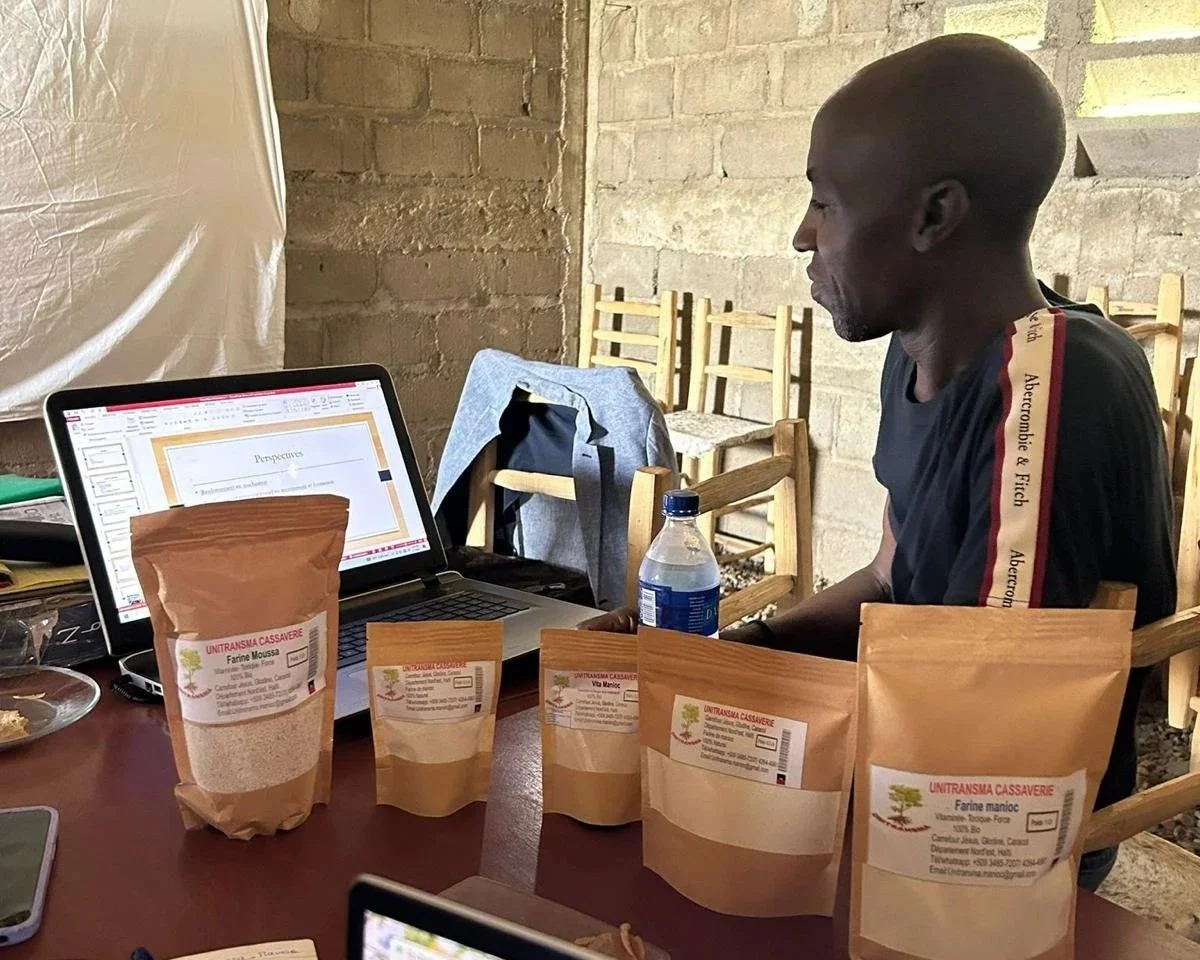 A man sitting at a table with a laptop, a water bottle, and several packages of flour, with a brick wall and empty chairs in the background.