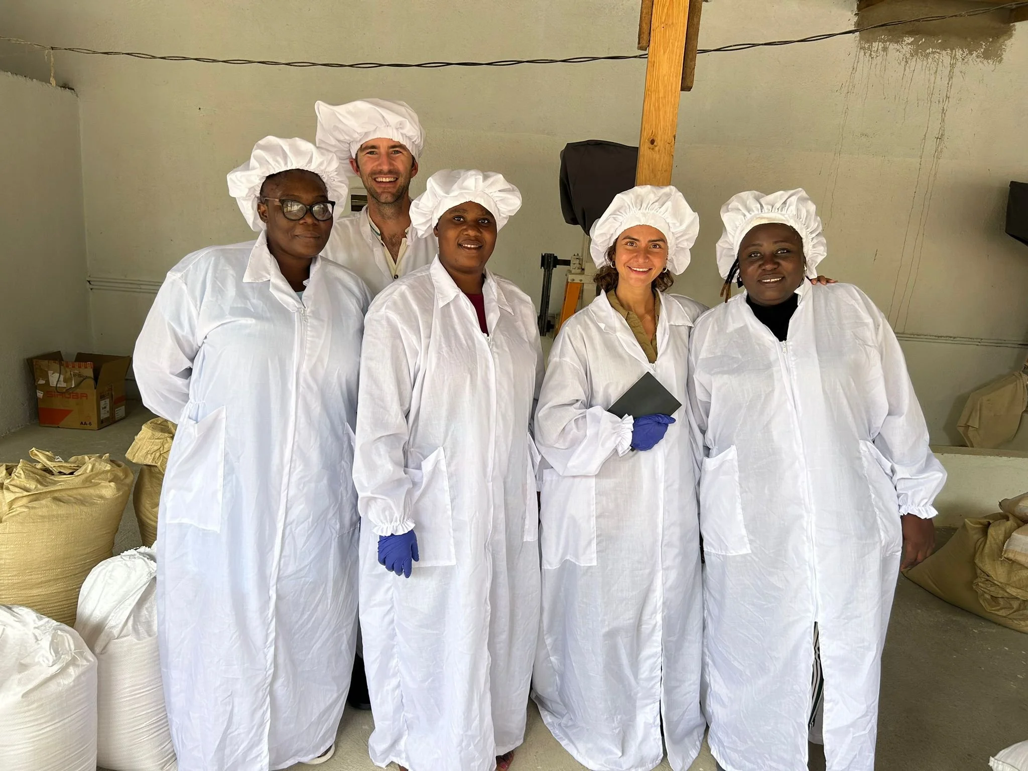 Six people wearing white protective suits and chef hats, standing together indoors, smiling at the camera.