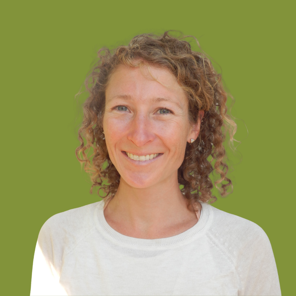 A woman with curly, light brown hair and earrings, smiling, wearing a white shirt, standing against a solid green background.