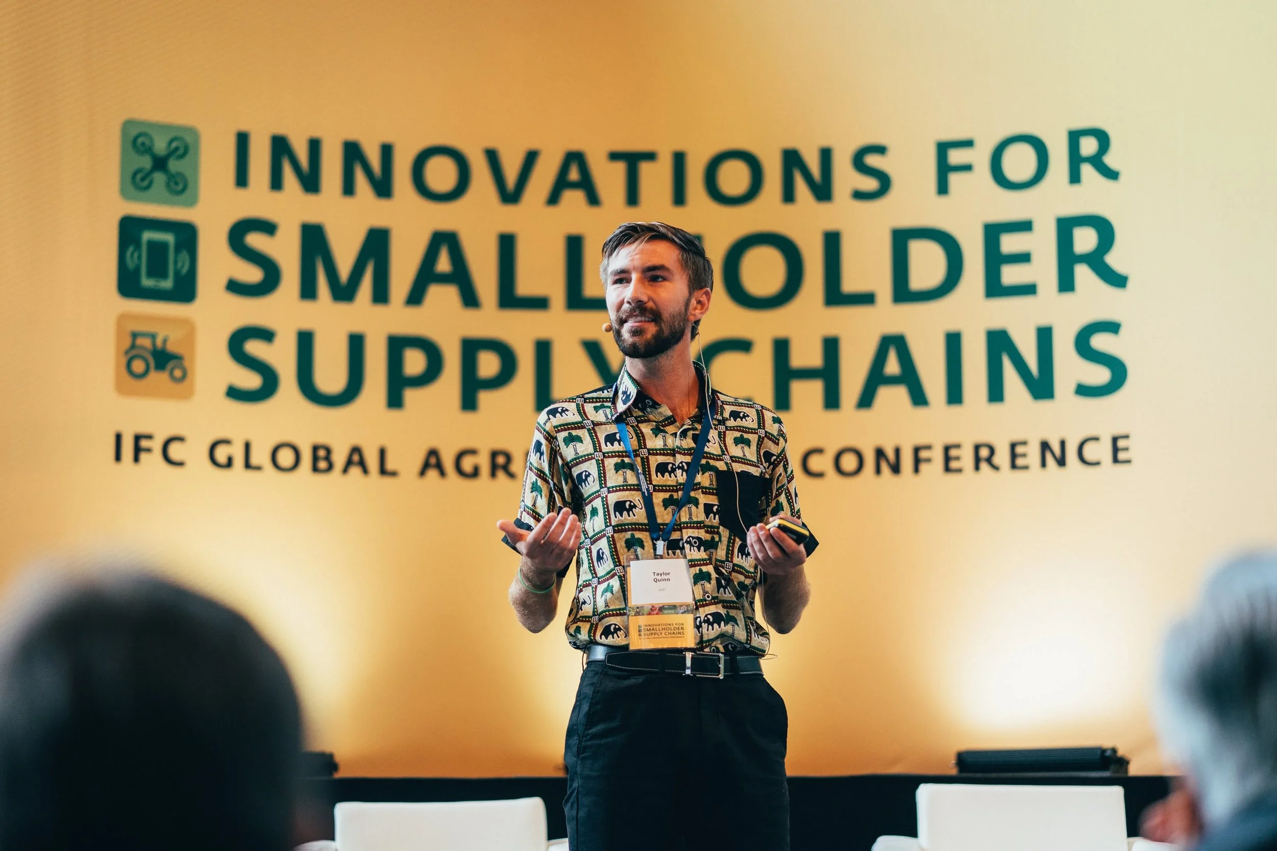 A man with a beard and patterned shirt is speaking at a conference with a large yellow banner behind him that reads 'Innovations for Smallholder Supply Chains'.