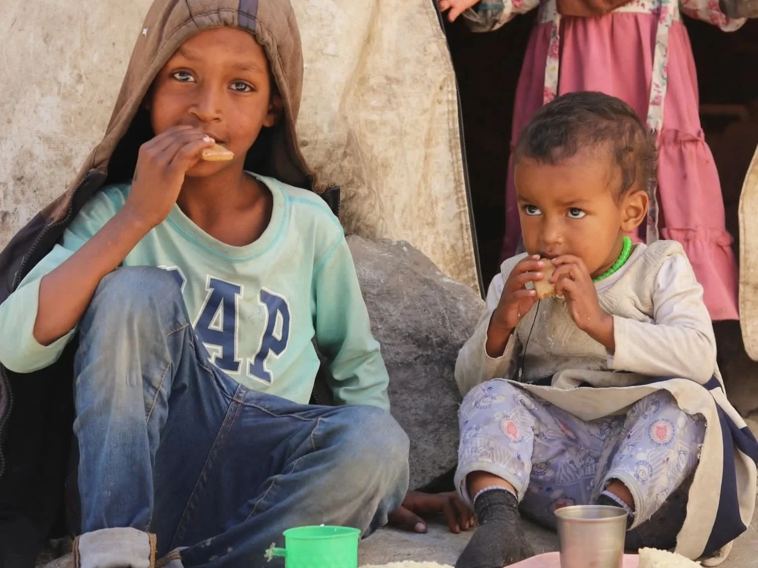 Two children with dark hair and skin sitting outdoors eating snacks.