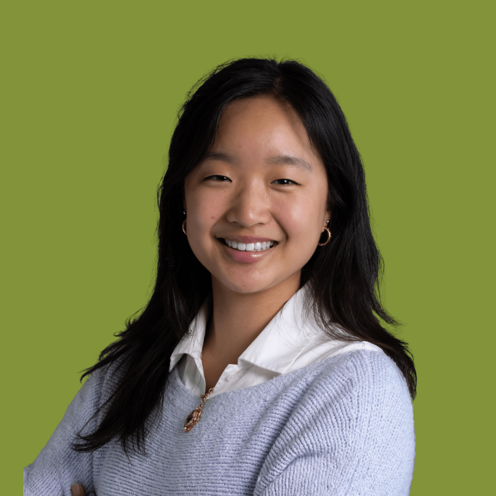 A young woman with long black hair smiling, wearing a white collared shirt under a light blue sweater, featuring hoop earrings and a necklace, against a green background.