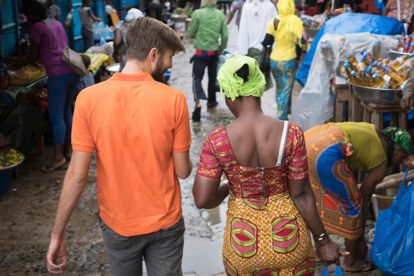 A man and a woman walking together through a crowded outdoor market on a rainy day. The man is wearing an orange shirt and gray pants, while the woman is dressed in colorful traditional African attire with a green headwrap. Surrounding them are various market stalls and other shoppers.