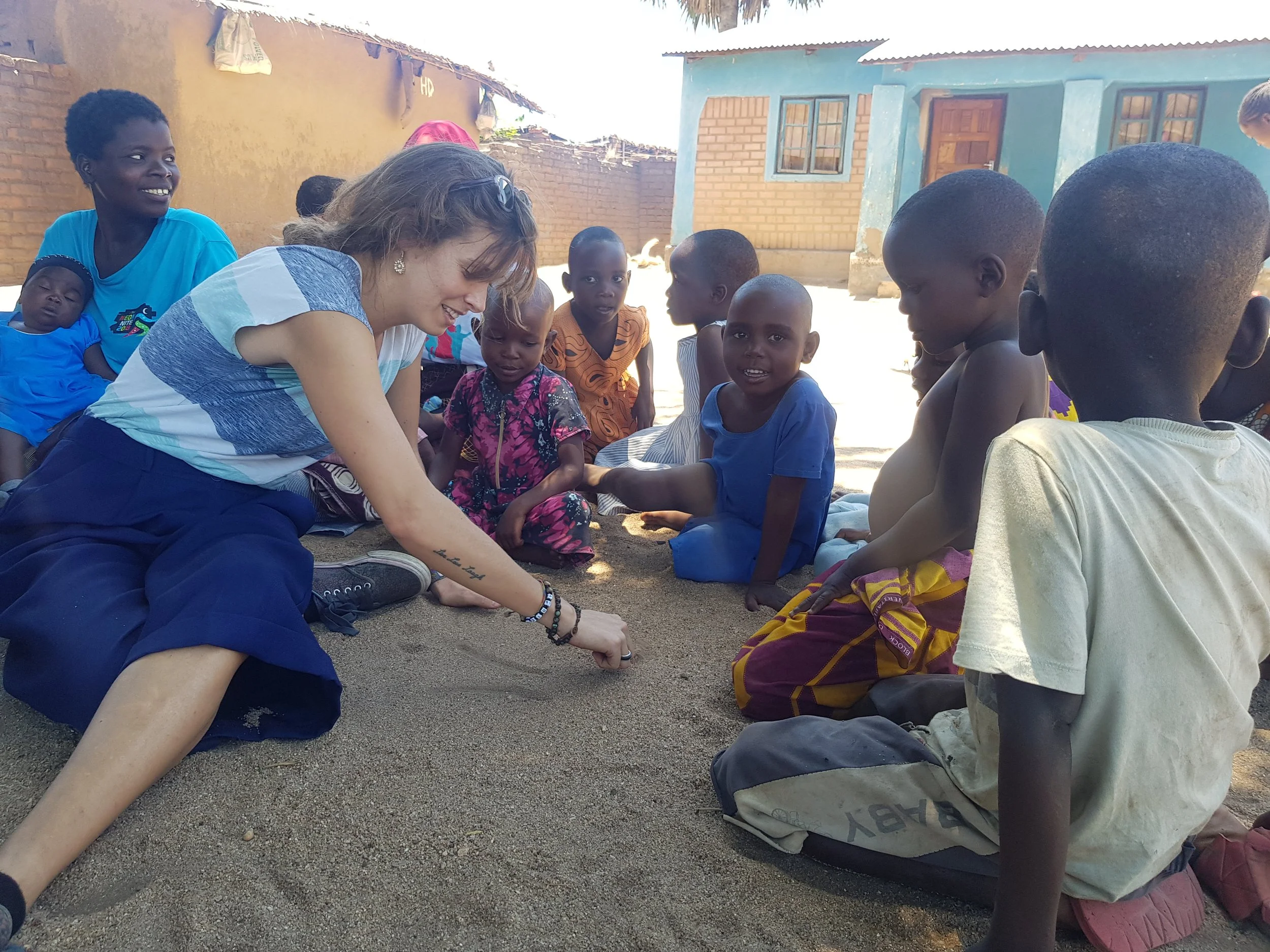 A group of children and a woman sitting outdoors, engaging in a discussion or activity. The setting appears to be in a rural area with simple houses in the background.