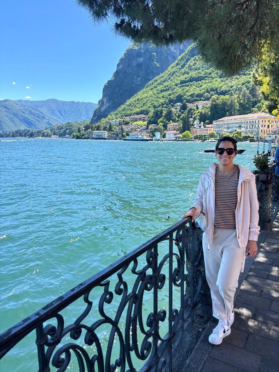 A woman wearing sunglasses, a light pink jacket, a striped shirt, and white pants stands by a metal railing along a lake, with mountains and buildings in the background under a clear blue sky.
