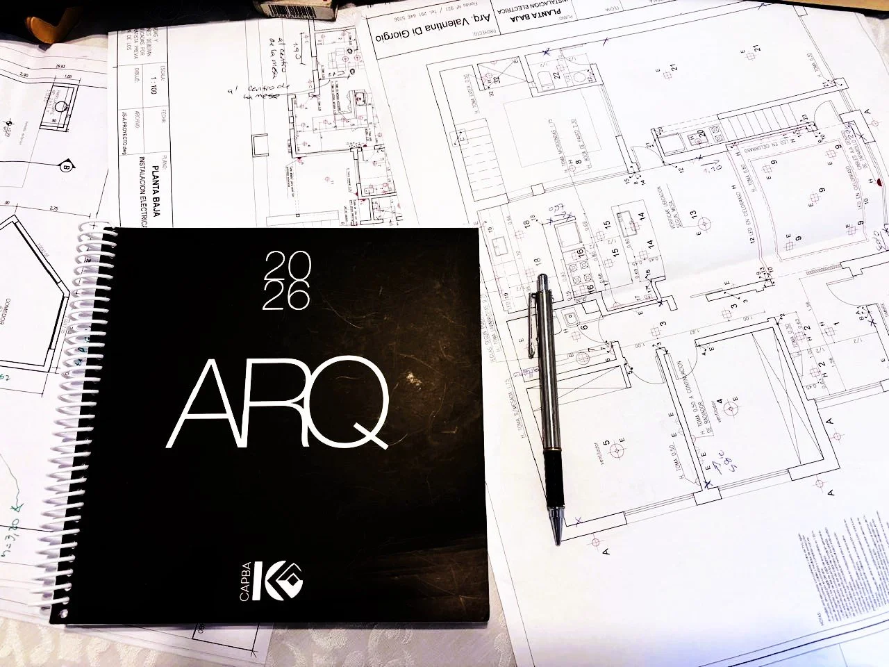 Architectural blueprints and a black designer notebook labeled 'ARQ 2026.'