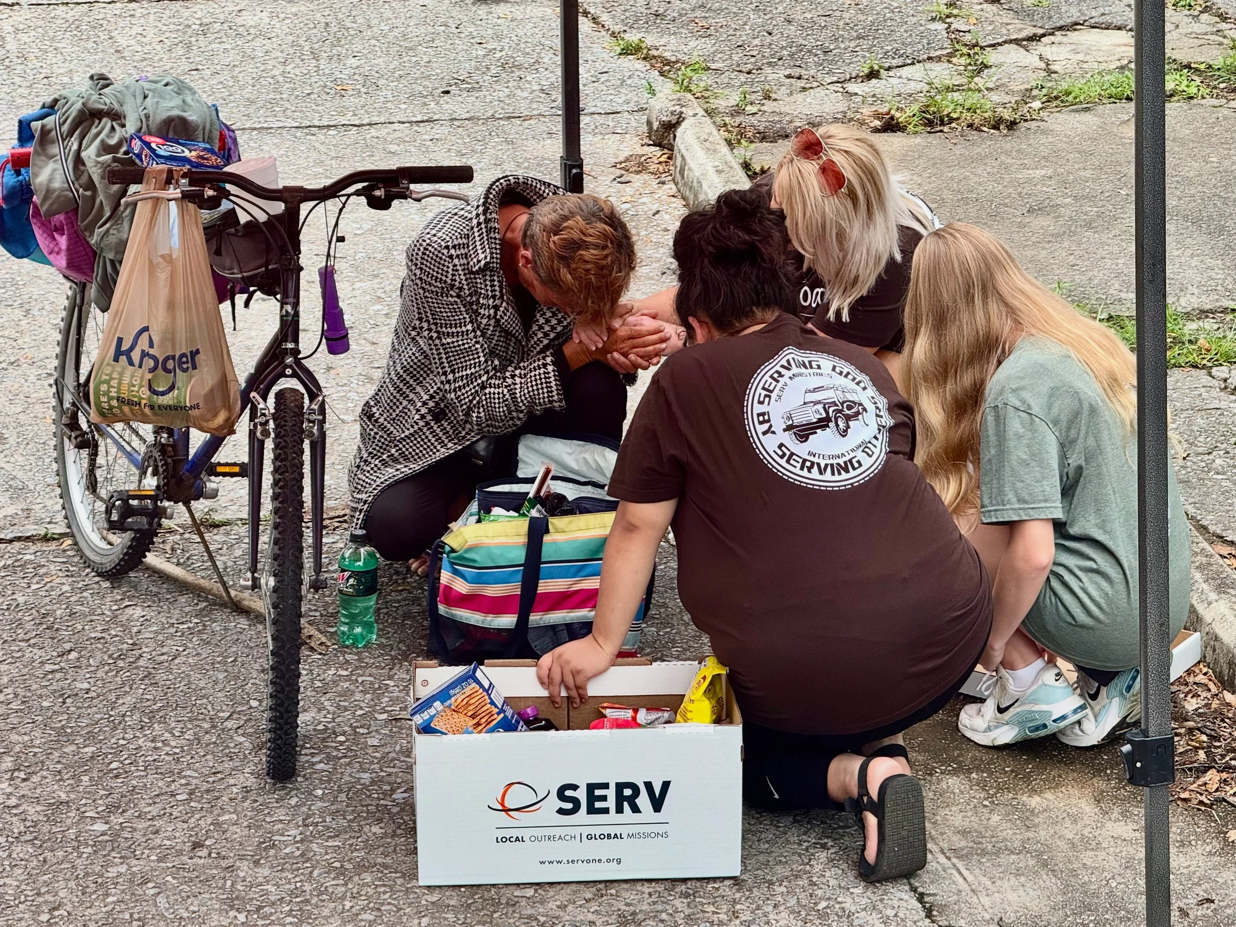 A group of people kneeling and sitting together at a charitable outreach event. They are exchanging or organizing food and supplies on a sidewalk. A bicycle with a bag of groceries is nearby, and a white box labeled 'SERV' with a website address is in front of them. The scene takes place outdoors on a paved surface with some patches of grass.