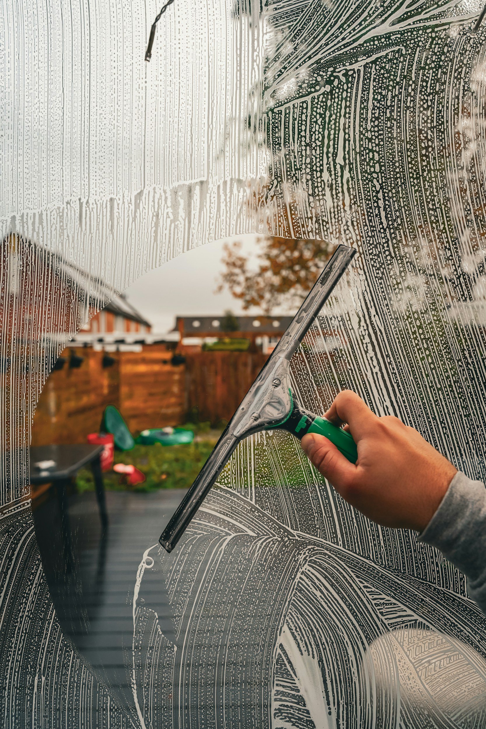 A person cleaning a glass window with a squeegee, with soap foam and water streaks on the glass. The background shows an outdoor backyard with a wooden fence, trees, and some outdoor furniture.
