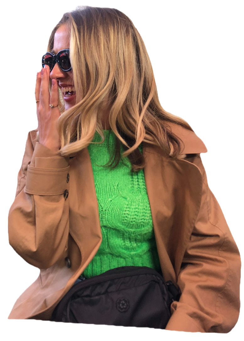 A woman laughing and covering her mouth with her hand, wearing sunglasses, a brown coat, a bright green sweater, and a black bag.