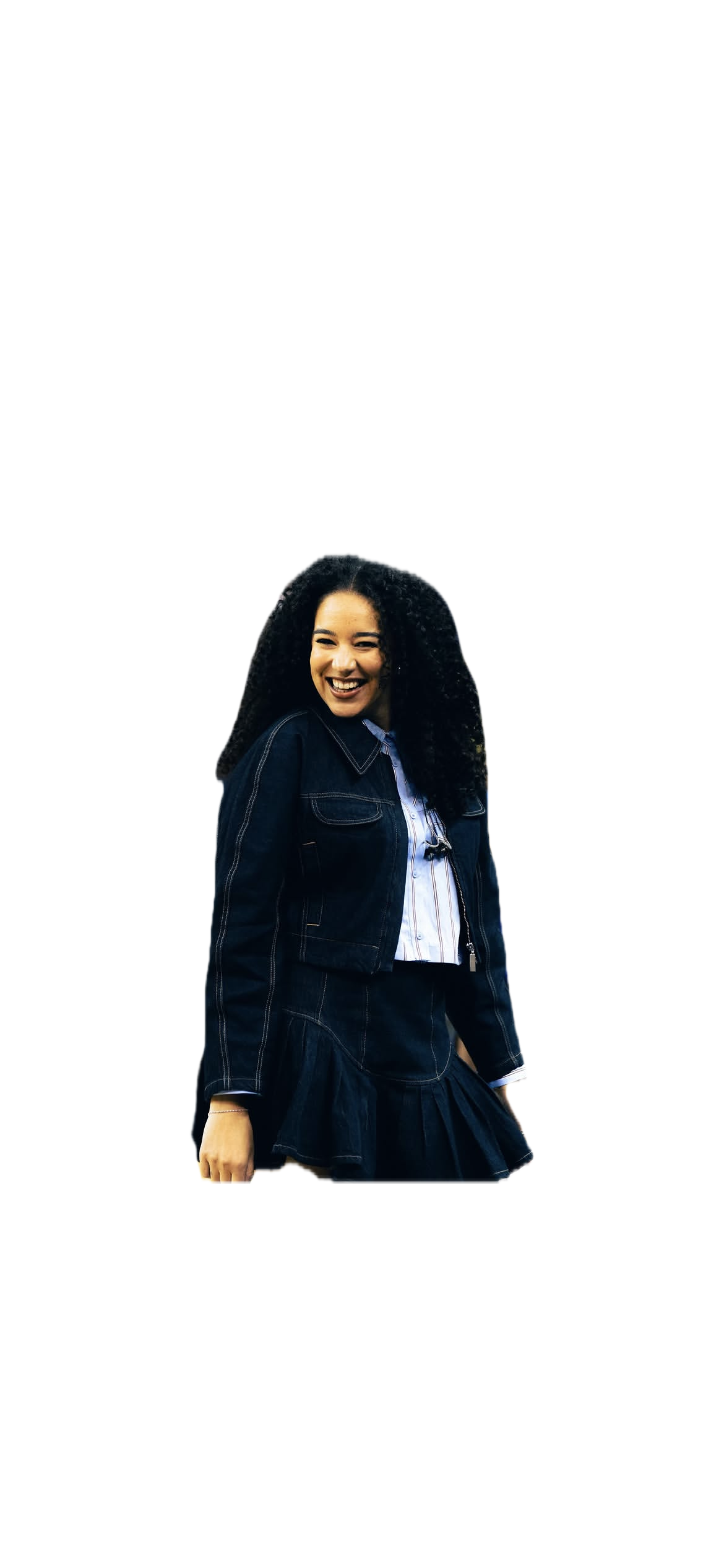 A young woman with curly black hair smiling, wearing a denim jacket and skirt, standing against a black background.