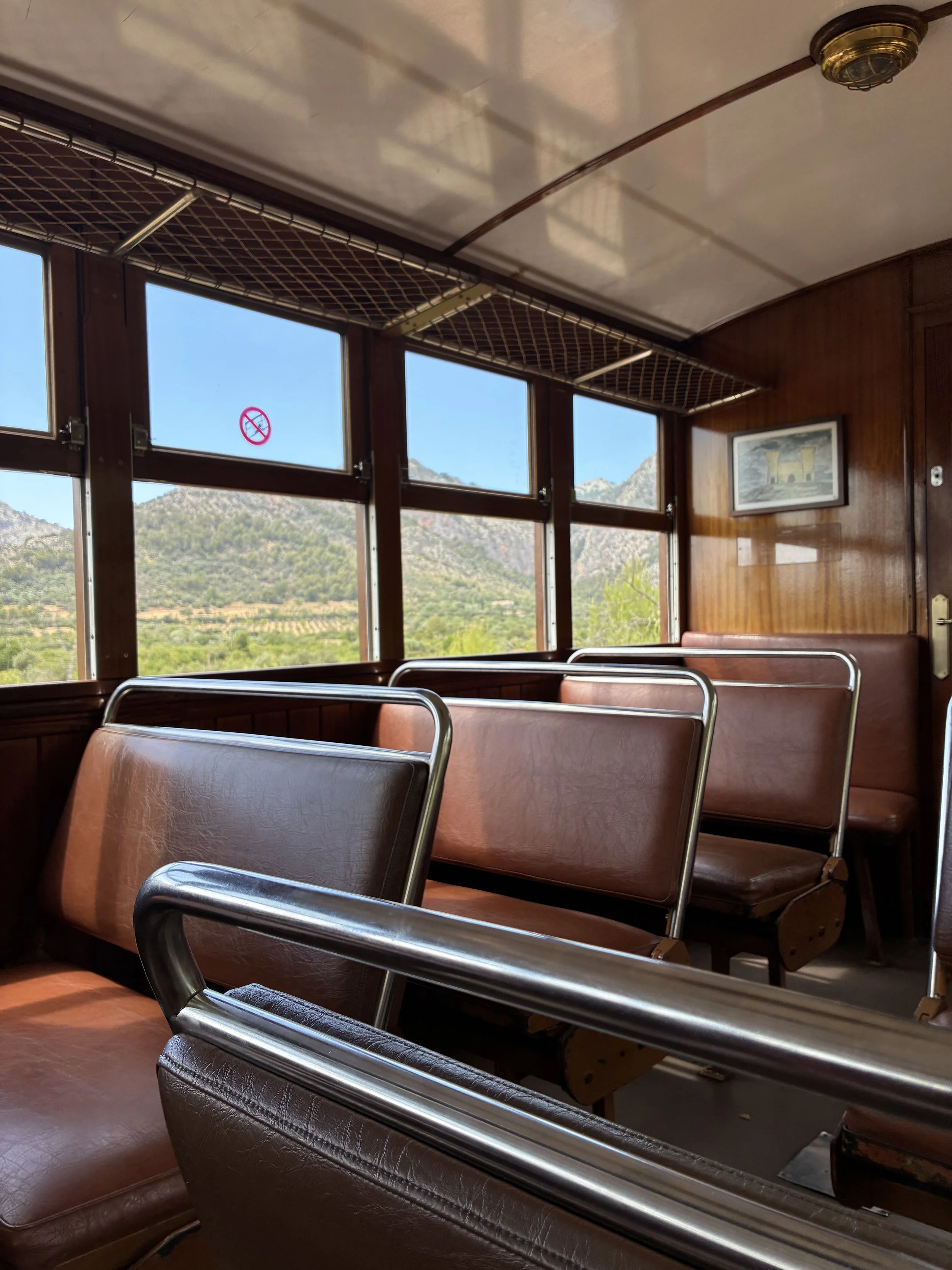 Empty vintage train car interior with brown leather seats, large windows showing mountains and landscape outside, framed picture on wall, and a no-smoking sign visible.
