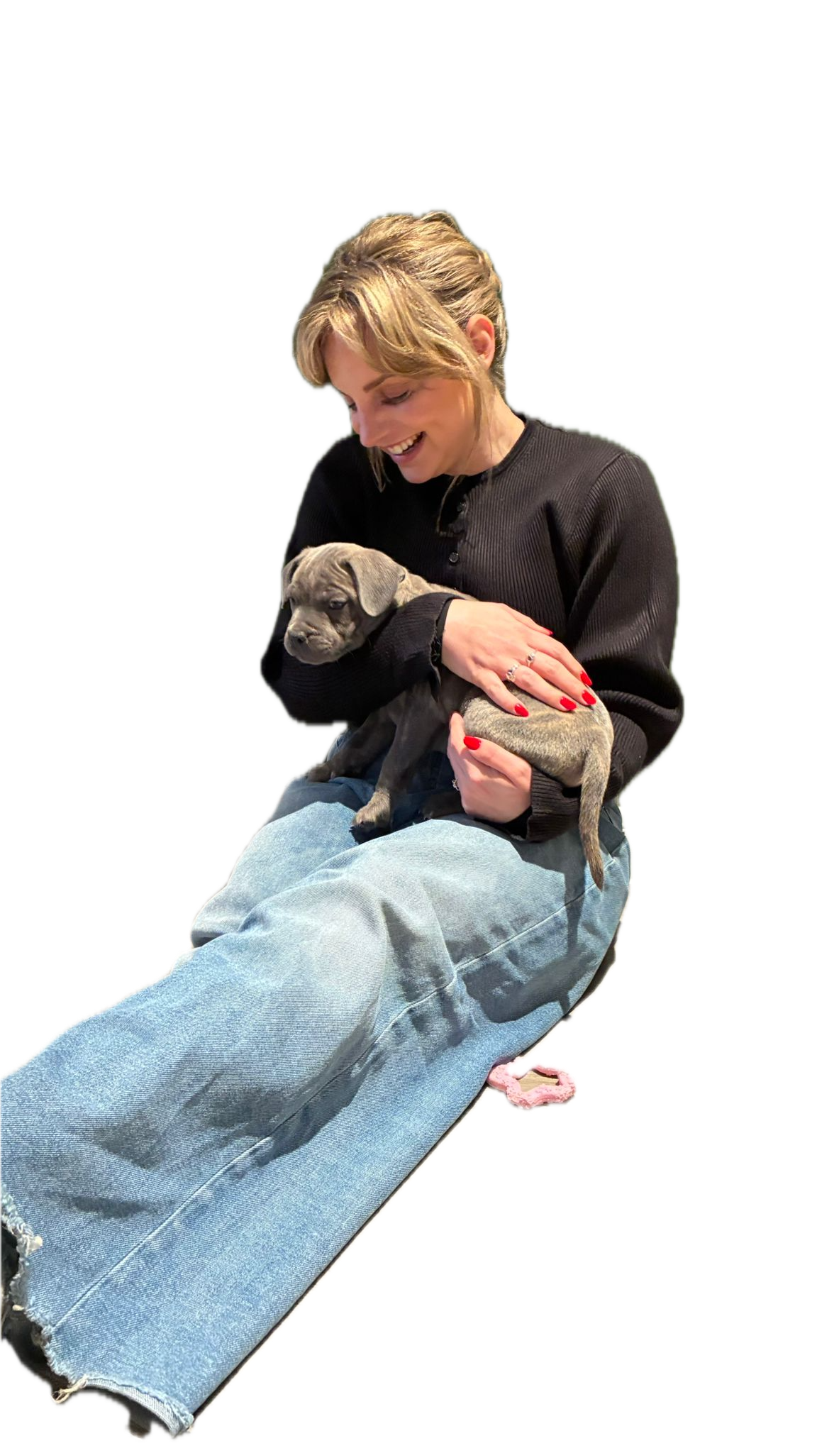 A woman with blonde hair, wearing a black top and light blue jeans, is sitting down and smiling while holding a small gray puppy. The puppy has a wrinkled face and floppy ears.