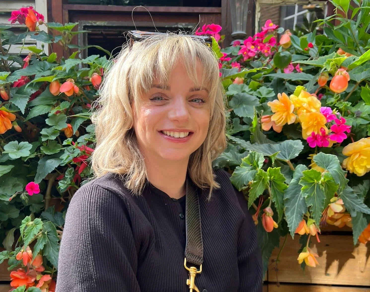 A woman with blonde hair smiling outdoors, standing in front of colorful pink, yellow, and orange flowers and green foliage.