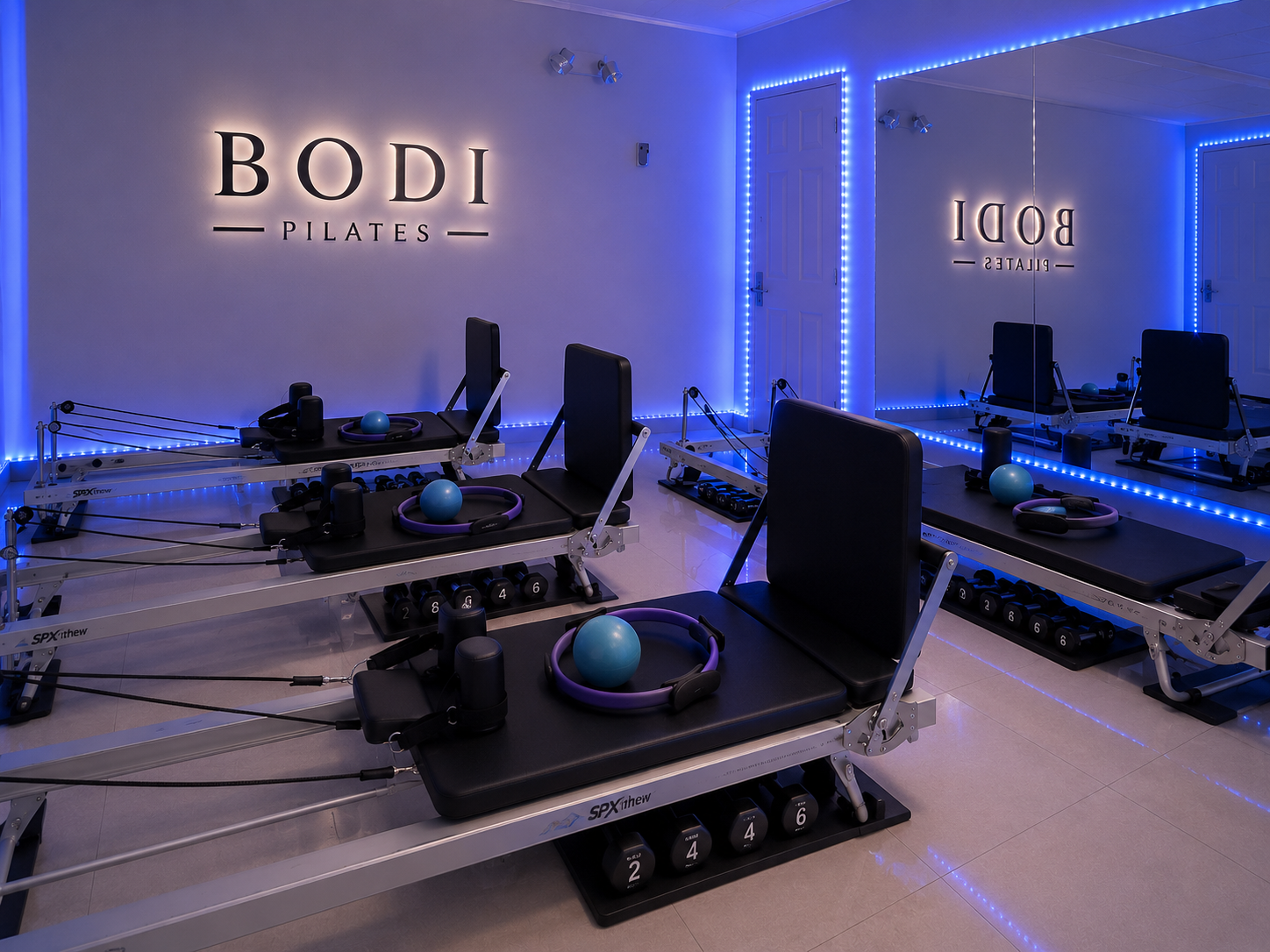 Pilates studio with reformer machines, blue lighting, and a large mirror on one wall, featuring the words 'BODI Pilates' illuminated on the wall.