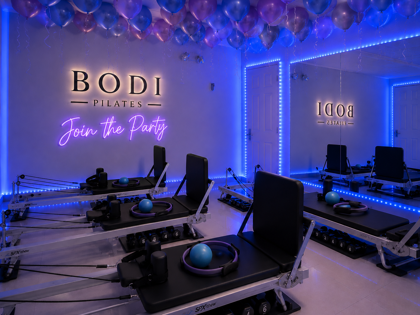 A Pilates studio decorated with blue and purple balloons on the ceiling, pink neon lights that read 'Join the Party', and a large mirror on the wall reflecting Pilates reformer machines with blue balls and resistance bands.