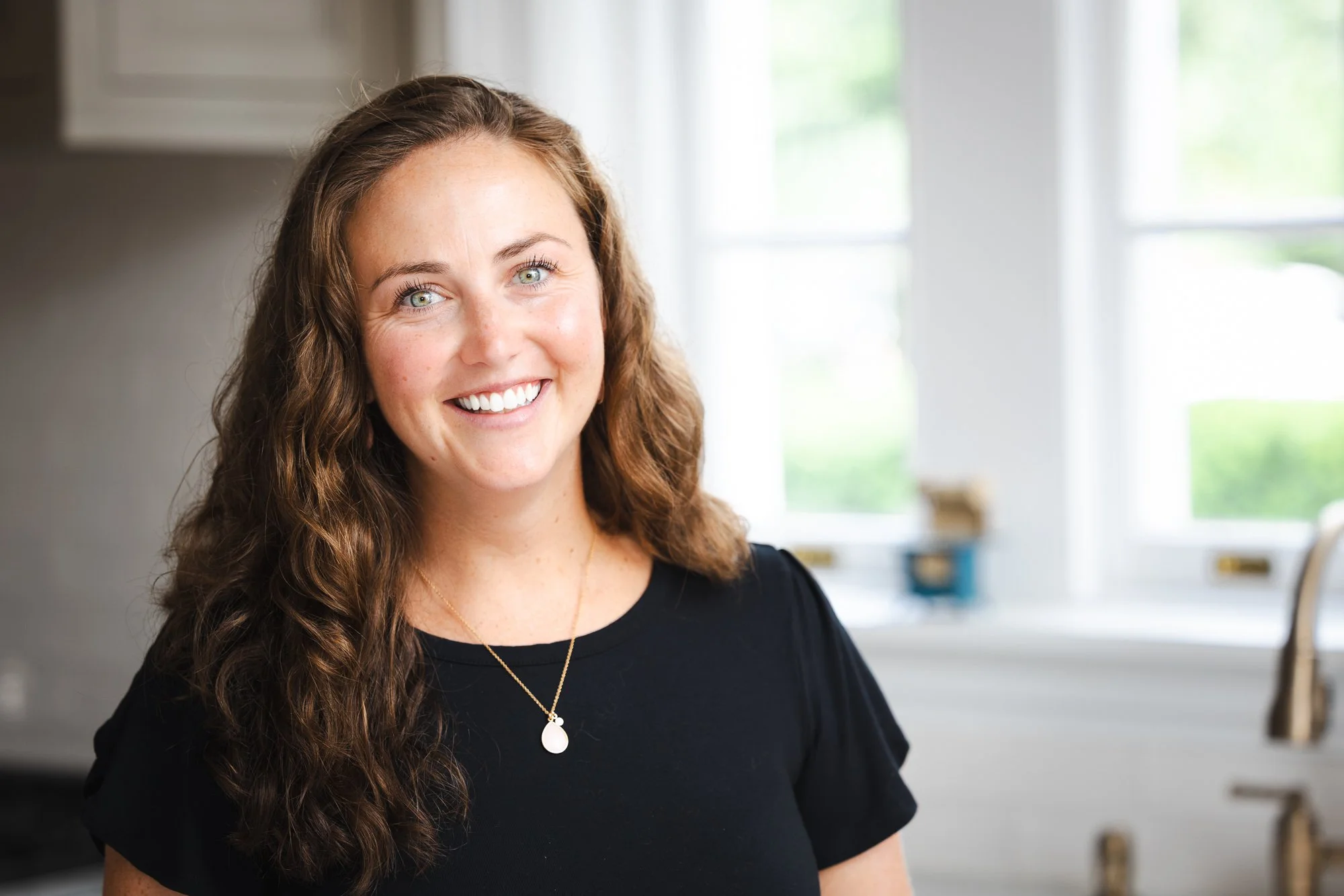 A woman with long curly brown hair, wearing a black top and gold necklace, smiling in a bright kitchen with large windows.