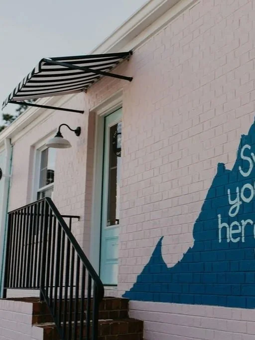 Exterior of a building with a black and white striped awning above a glass door, pink brick wall, a black metal staircase, and a blue mural with white text.