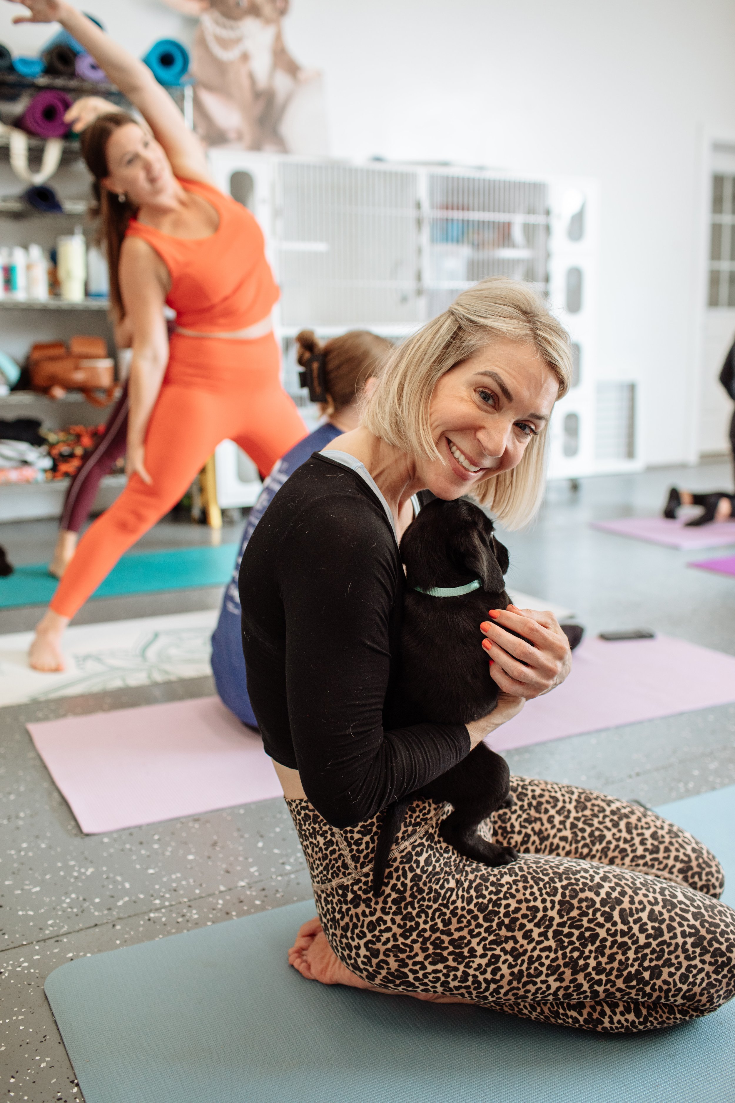 A woman with blonde hair sitting on a yoga mat, hugging a black puppy, in a yoga or fitness studio. In the background, other women are exercising, with one woman stretching in an orange outfit.