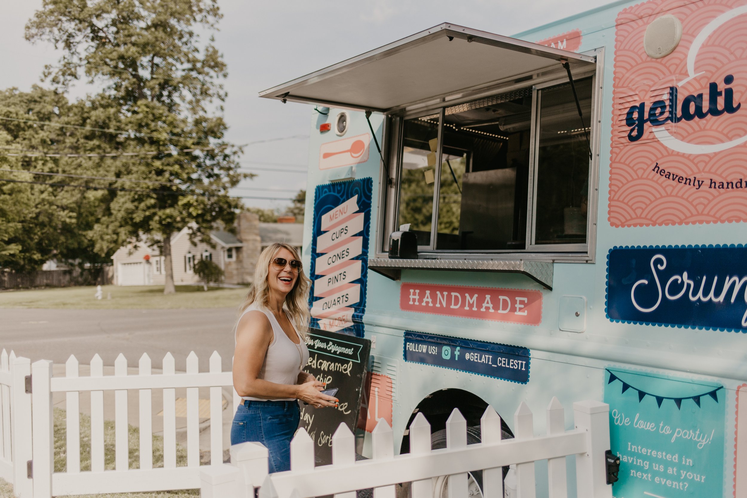 A woman with blonde hair and sunglasses smiling standing in front of a pink and blue gelato food truck, holding her phone. The truck has menu signs and social media info, and is parked in a grassy area with houses and trees in the background, separated by a white picket fence.