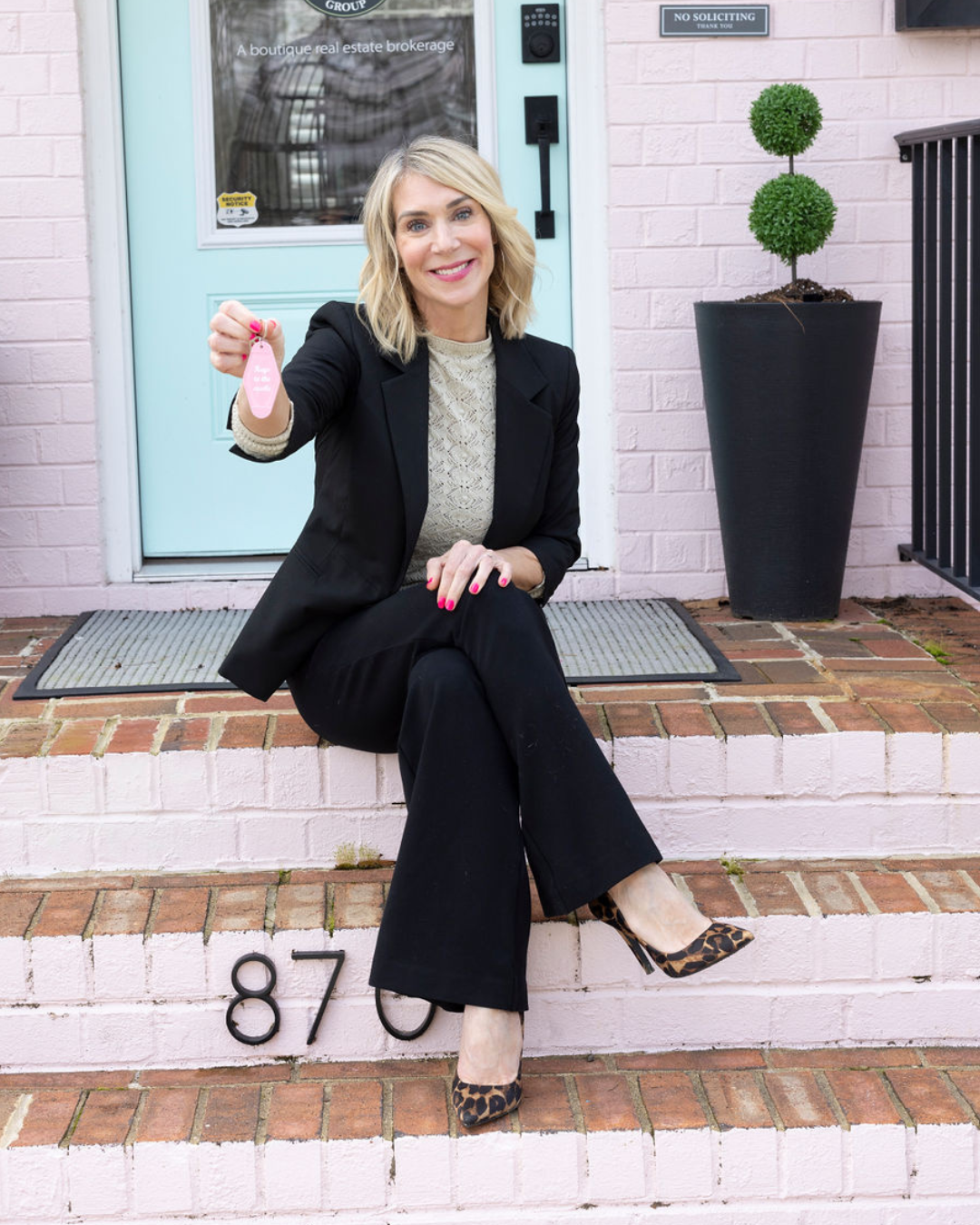 Woman in business attire sitting on brick stairs in front of a teal door, holding a pink keychain; house number 870 on stairs; potted plant beside door.