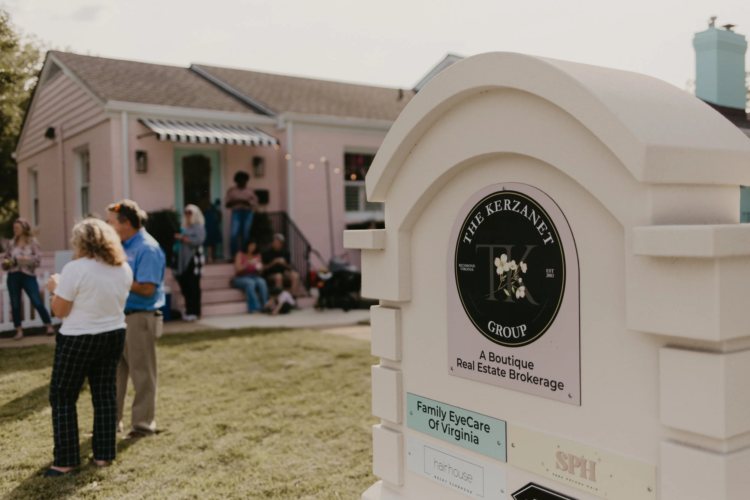 People gathering outside a pink house with a striped awning, and a prominent white sign with black and gold text for The Kerzane Group real estate brokerage in Bon Air, Virginia.