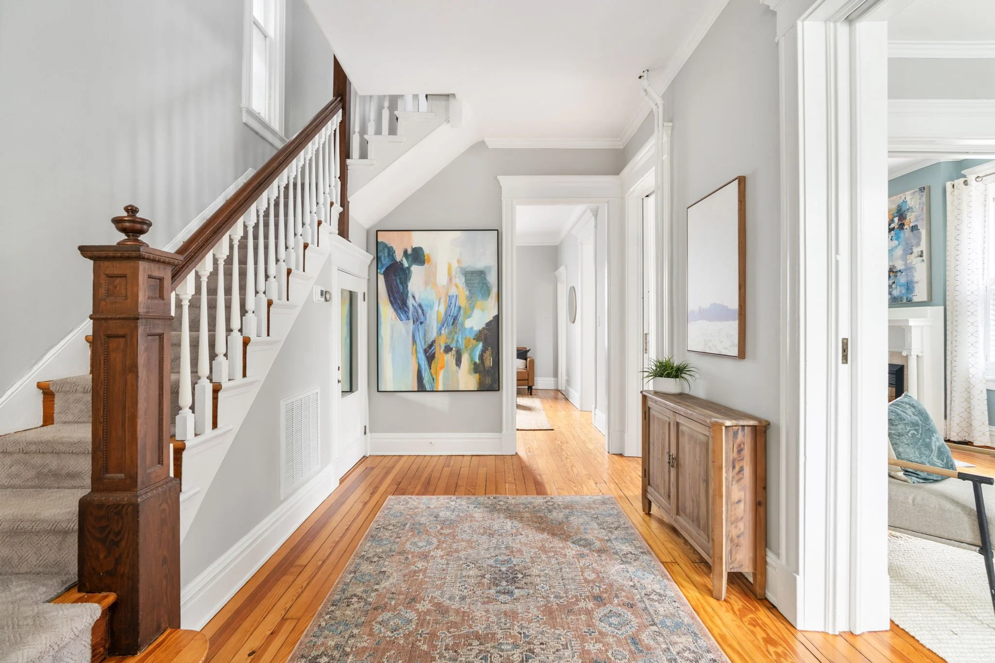 A welcoming hallway in a Richmond, Virginia luxury home with hardwood floors, a patterned rug, white walls, and artwork hanging. Stairs with a wooden banister lead to an upper floor, and natural light fills the space.