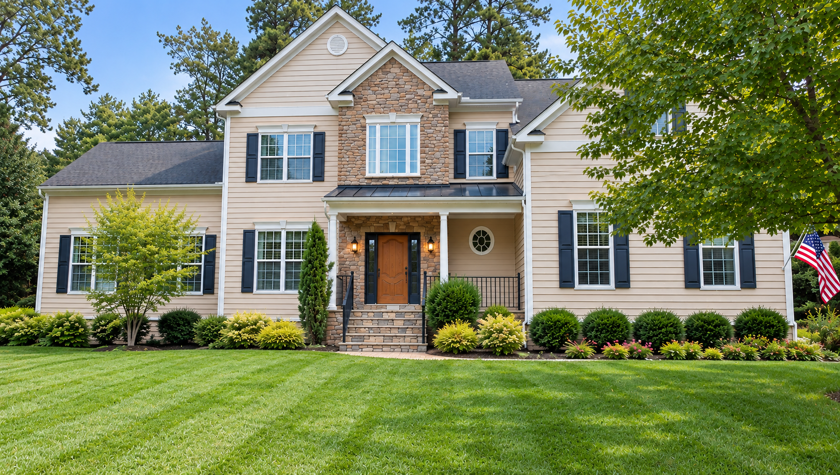 A two-story suburban house with beige siding, dark shutters, and a brick and stone facade, surrounded by a well-kept lawn and landscaped bushes and trees.