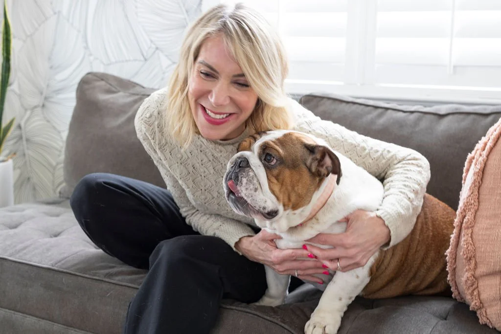 A woman smiling and cuddling with a bulldog on a gray sofa in a well-lit living room.