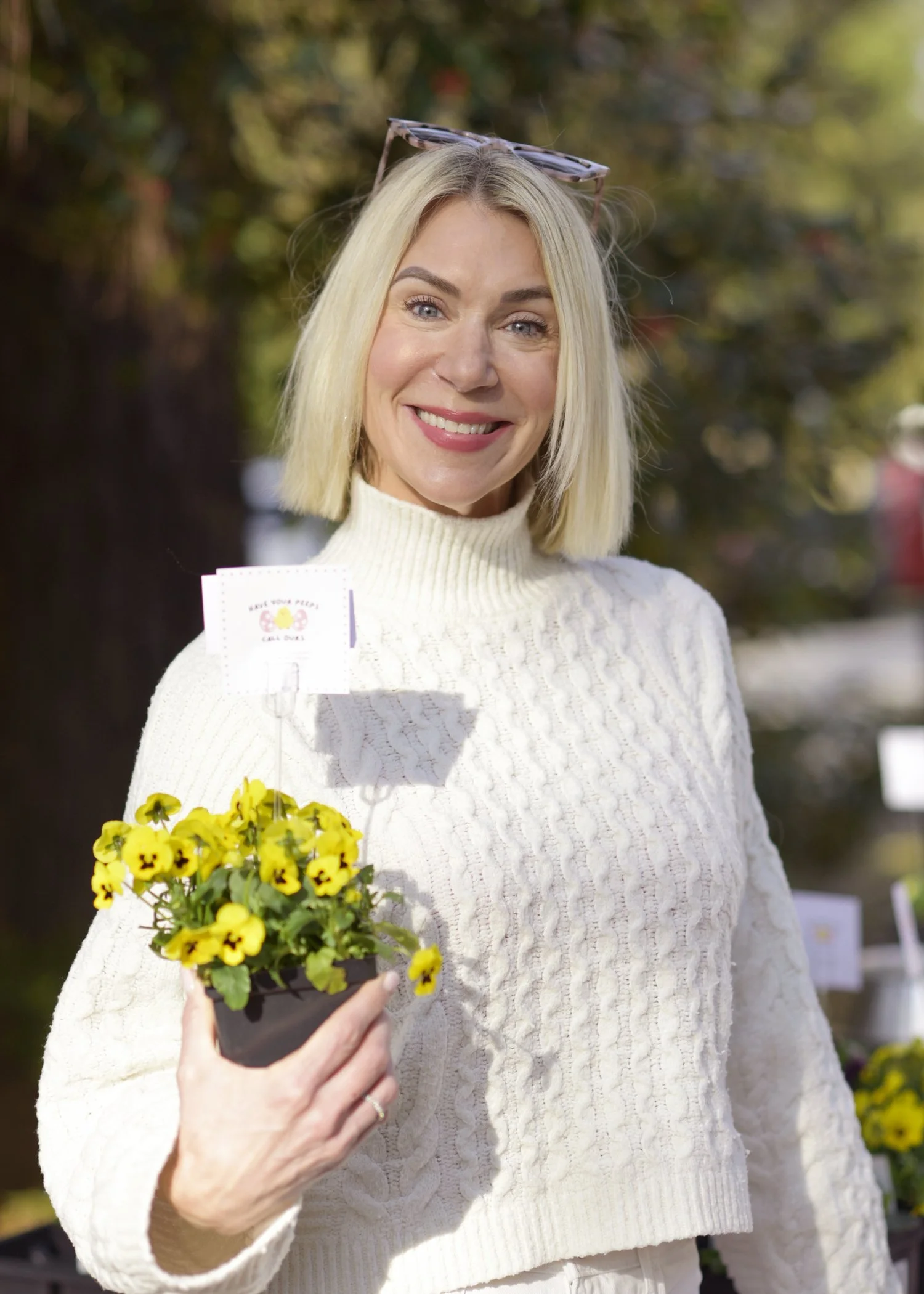 A woman with blonde hair wearing a white sweater, holding a small pot of yellow flowers in front of her, smiling outdoors with trees in the background.