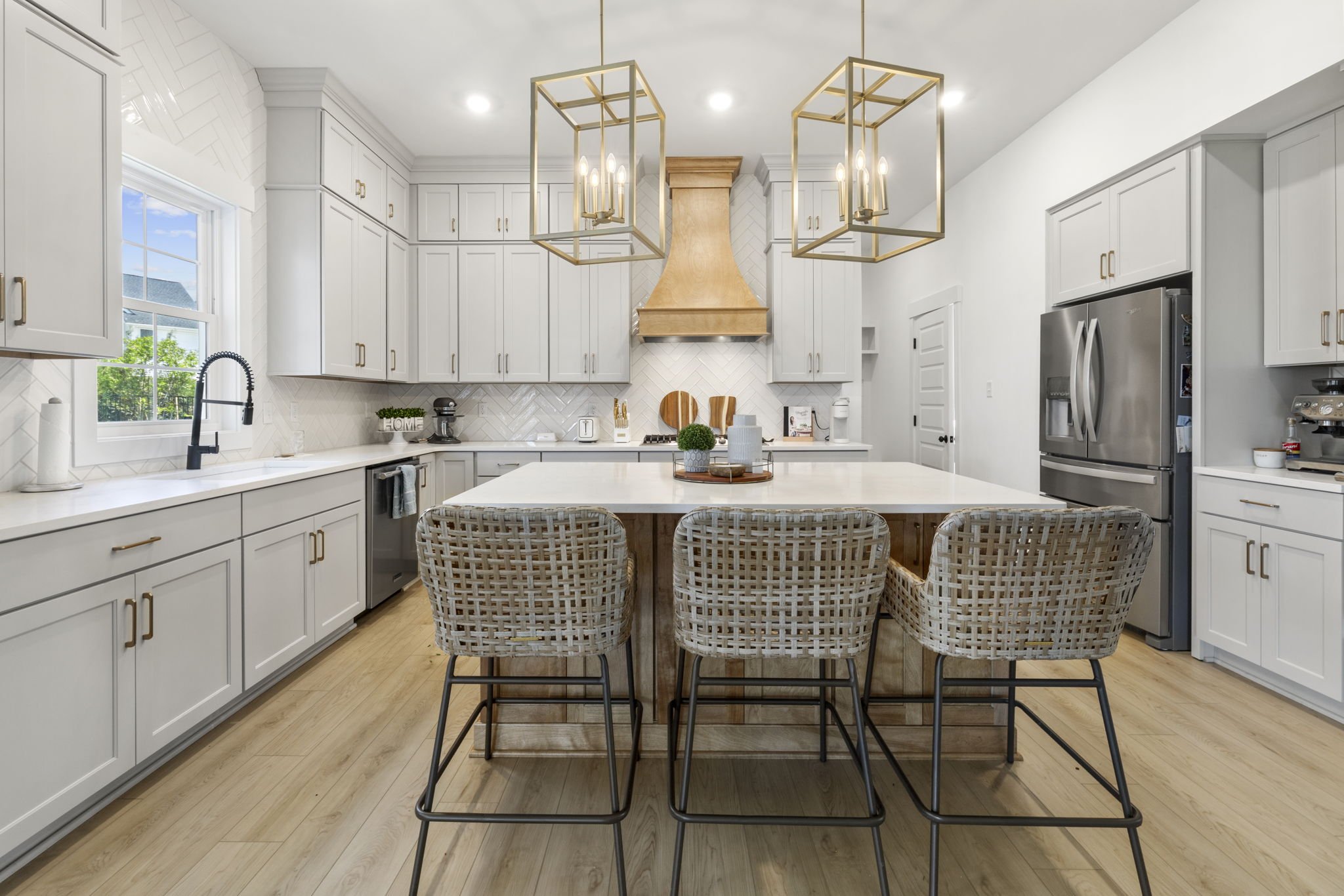 Modern kitchen with white cabinets, a white island, black faucet, stainless steel refrigerator, and hanging gold light fixtures over the island. Wood flooring and a window showing greenery outside.