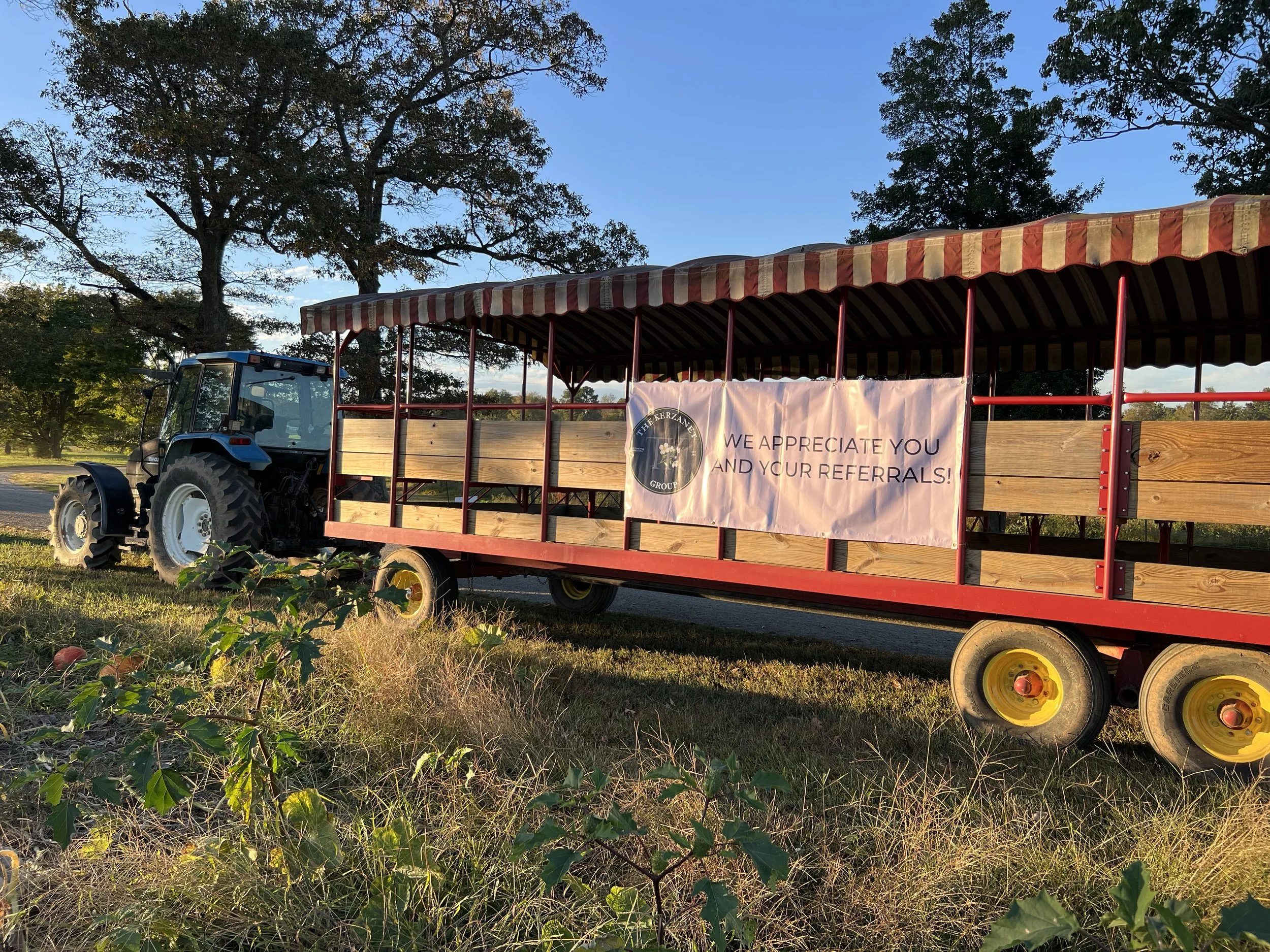 A tractor with a red and yellow striped canopy attached to a wooden platform with a Kerzanet Groupbanner that reads, 'We appreciate you and your referrals.' The tractor is parked outdoors near trees and grass, with a clear blue sky in the background.