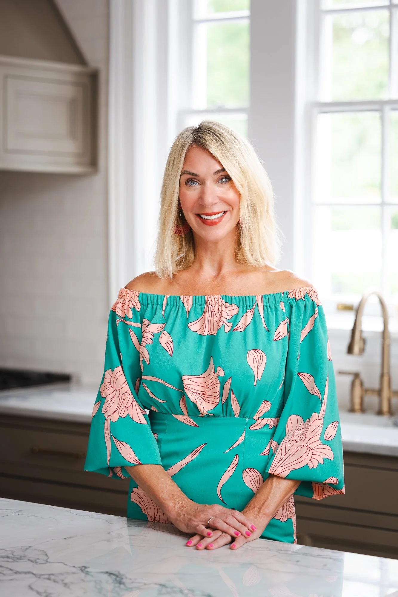 A smiling woman with blonde hair, wearing a teal off-the-shoulder dress with pink floral patterns, standing in a bright kitchen near a marble counter.