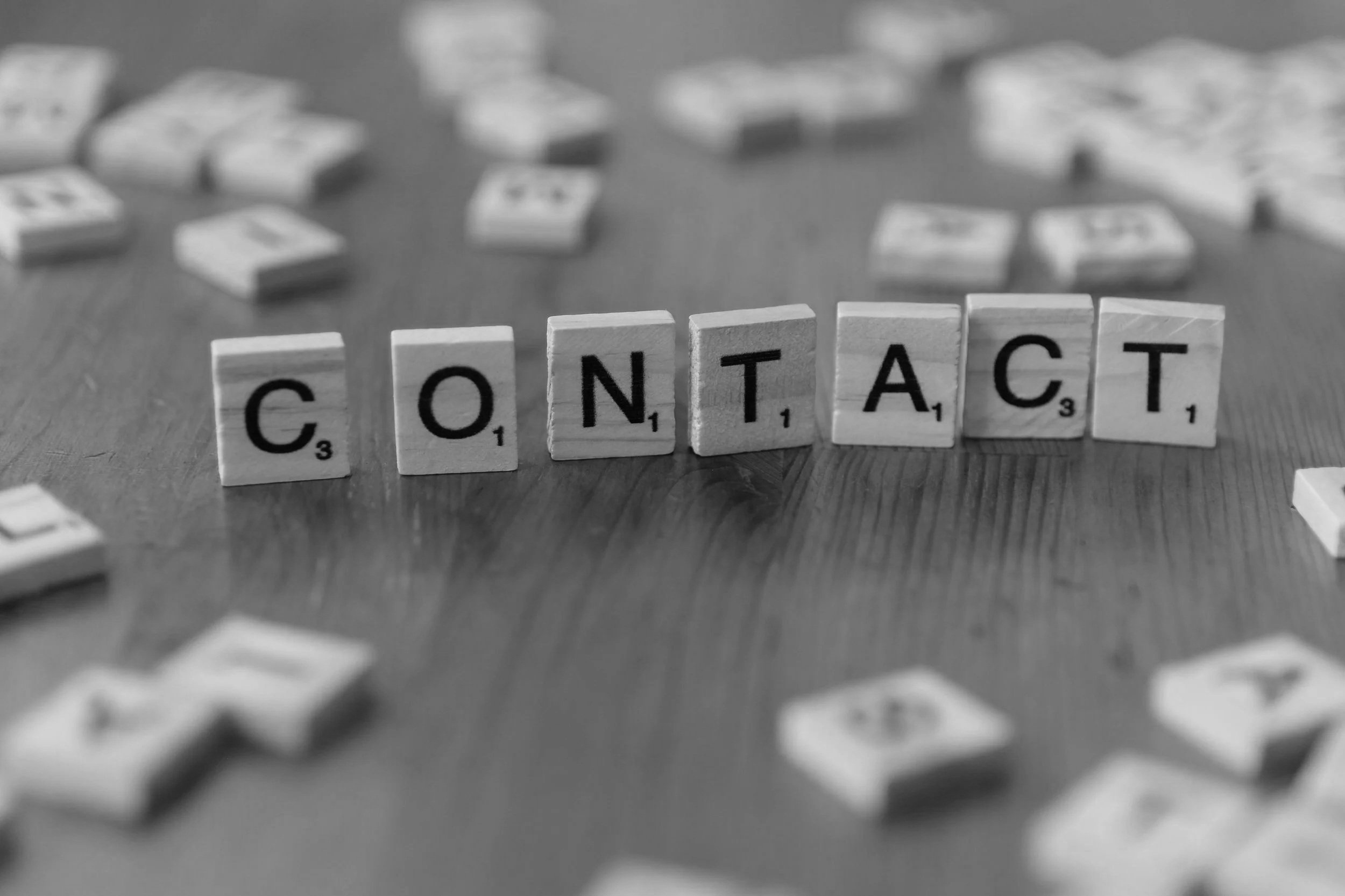Black and white photograph of wooden Scrabble tiles spelling the word 'CONTACT' on a table, with scattered tiles and blurred game pieces in the background.