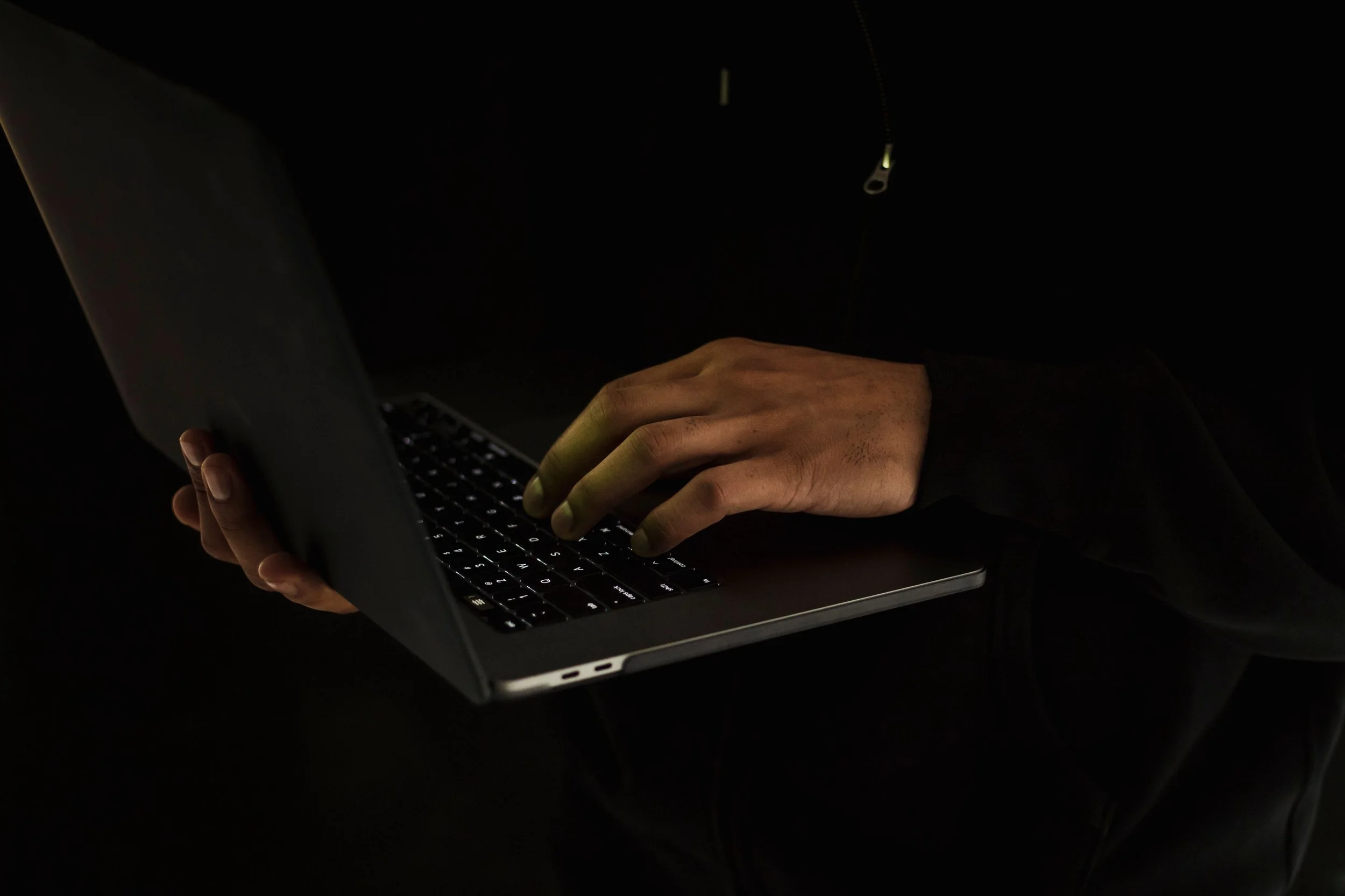 A person typing on a laptop keyboard in a dark environment, with only the keyboard and hands illuminated.