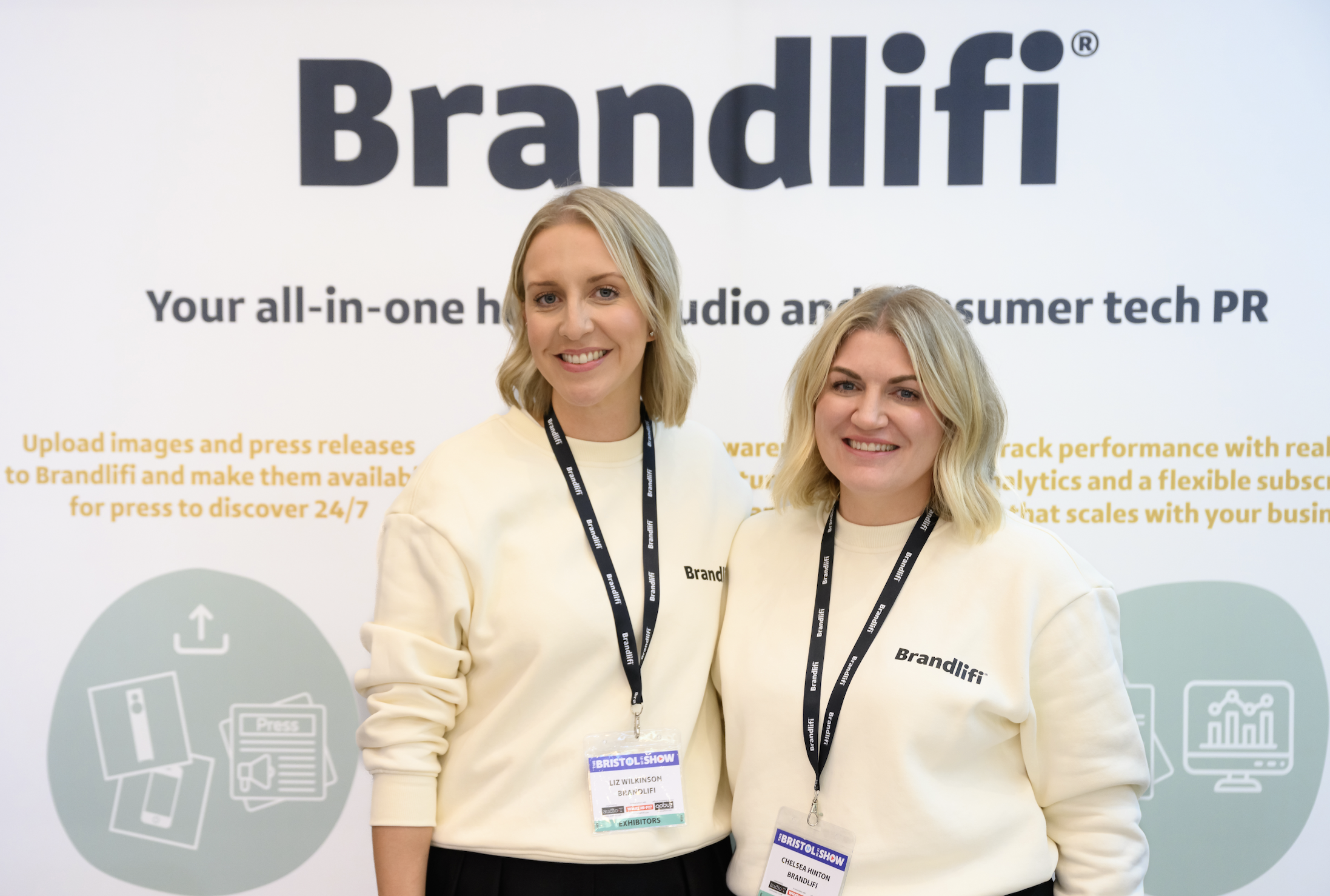 Chelsea Hinton and Liz Wilkinson standing in front of a white backdrop with branding and icons, both wearing light-colored sweaters and black lanyards with tags, smiling at the camera.