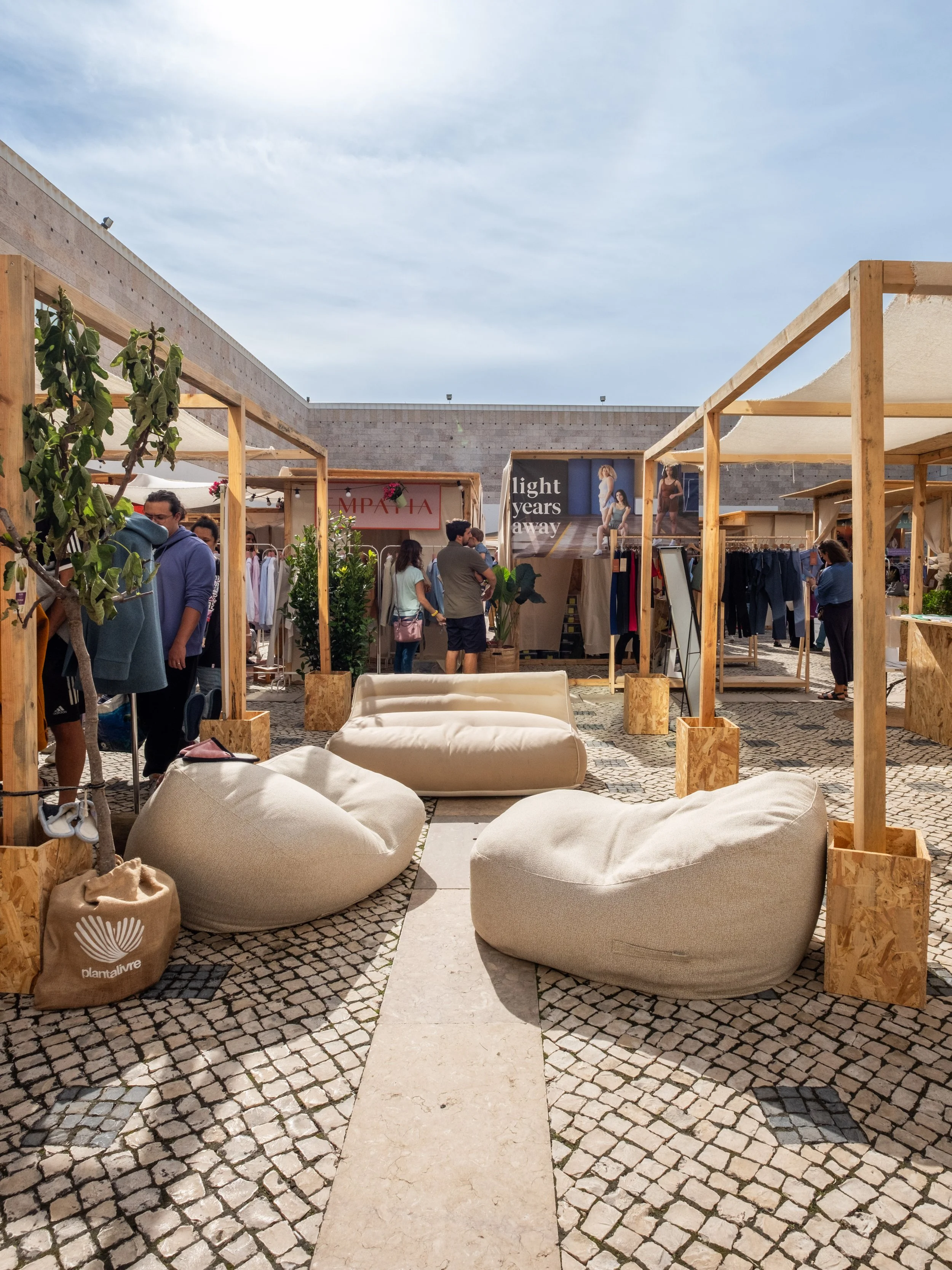 An outdoor marketplace with wooden stalls, people browsing, and large bean bag chairs on a stone-paved surface, under a clear sky.