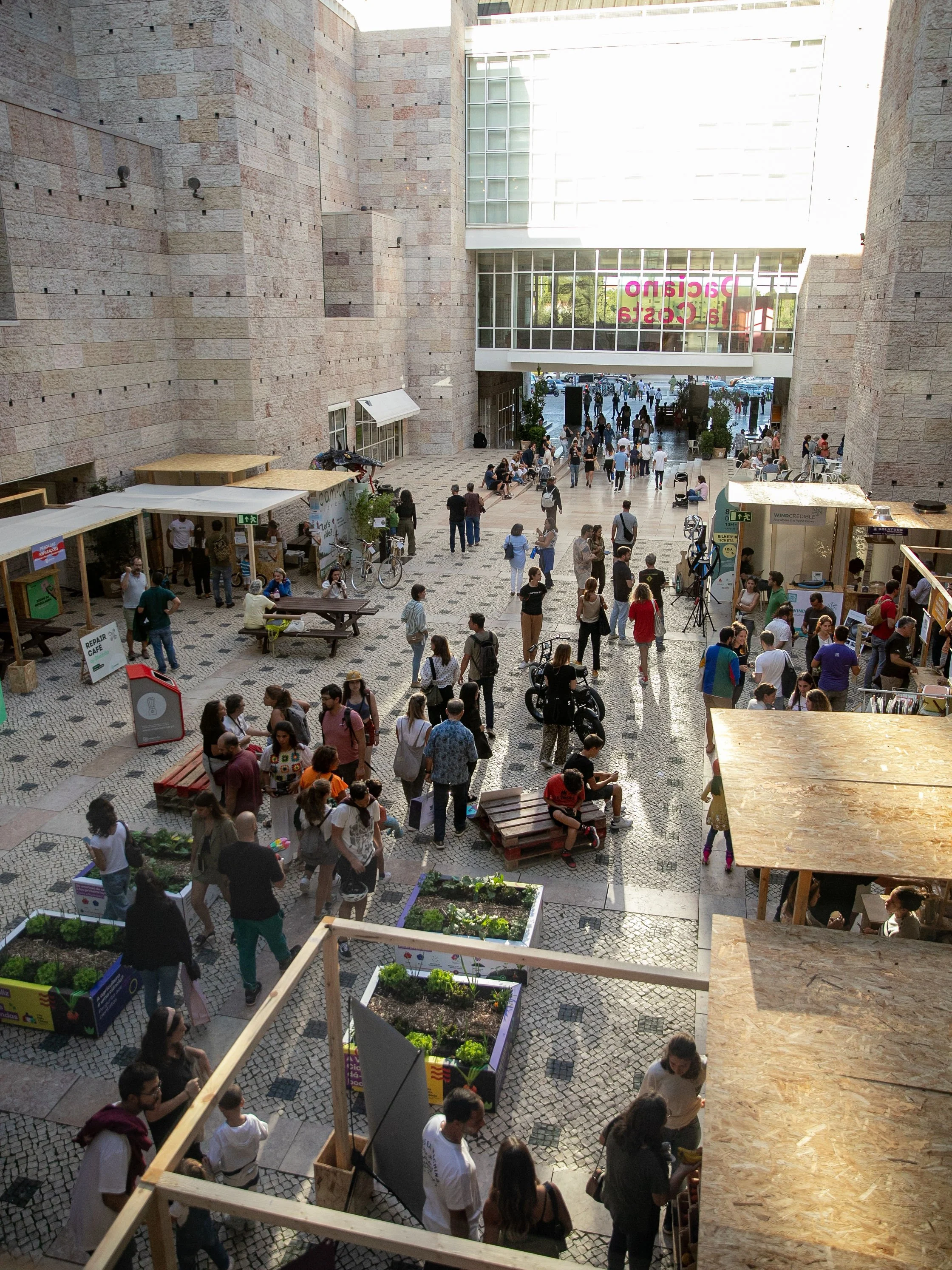 People gather at an outdoor market or event in a courtyard area inside a large modern building with brick walls and large glass windows. There are small stalls, seating areas, and planters with vegetables, and some people are walking, talking, and browsing.