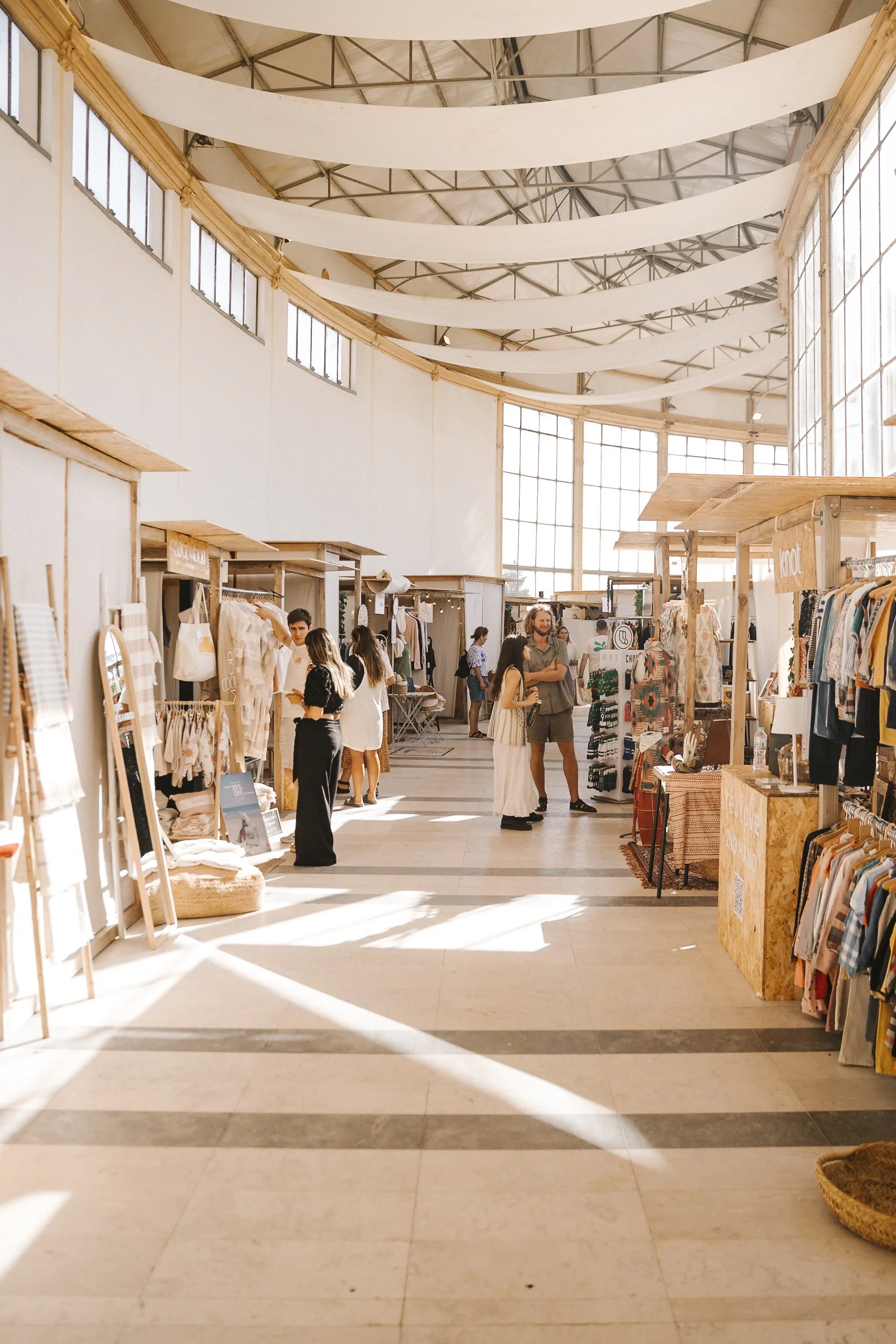 Interior of a spacious marketplace or craft fair with vendors displaying clothing and accessories. Sunlight streams through large windows, illuminating the scene with shoppers browsing and chatting.