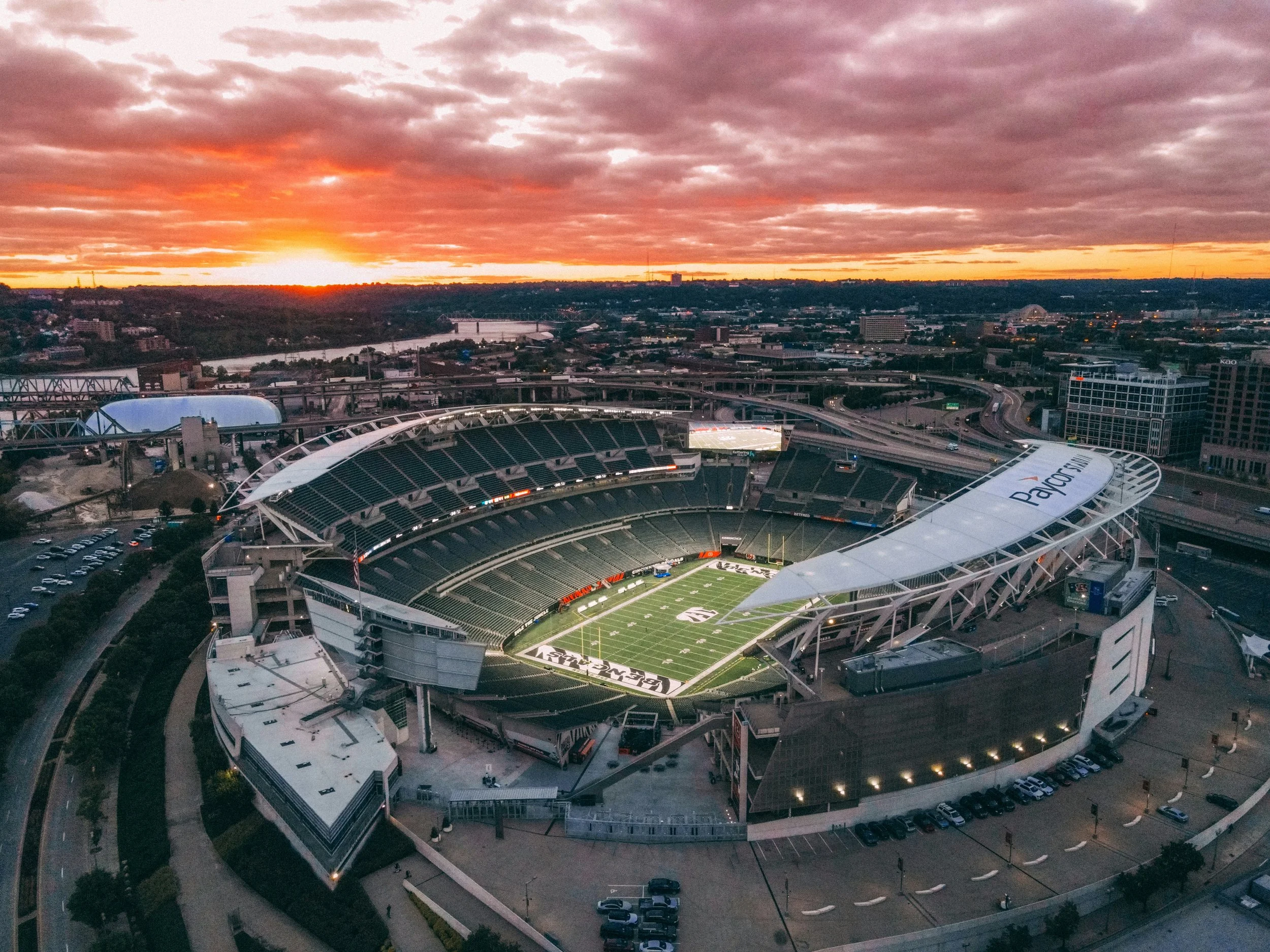 Aerial view of Netflix Stadium at sunset, with orange and purple clouds, the football field inside the stadium, and cityscape in the distance.
