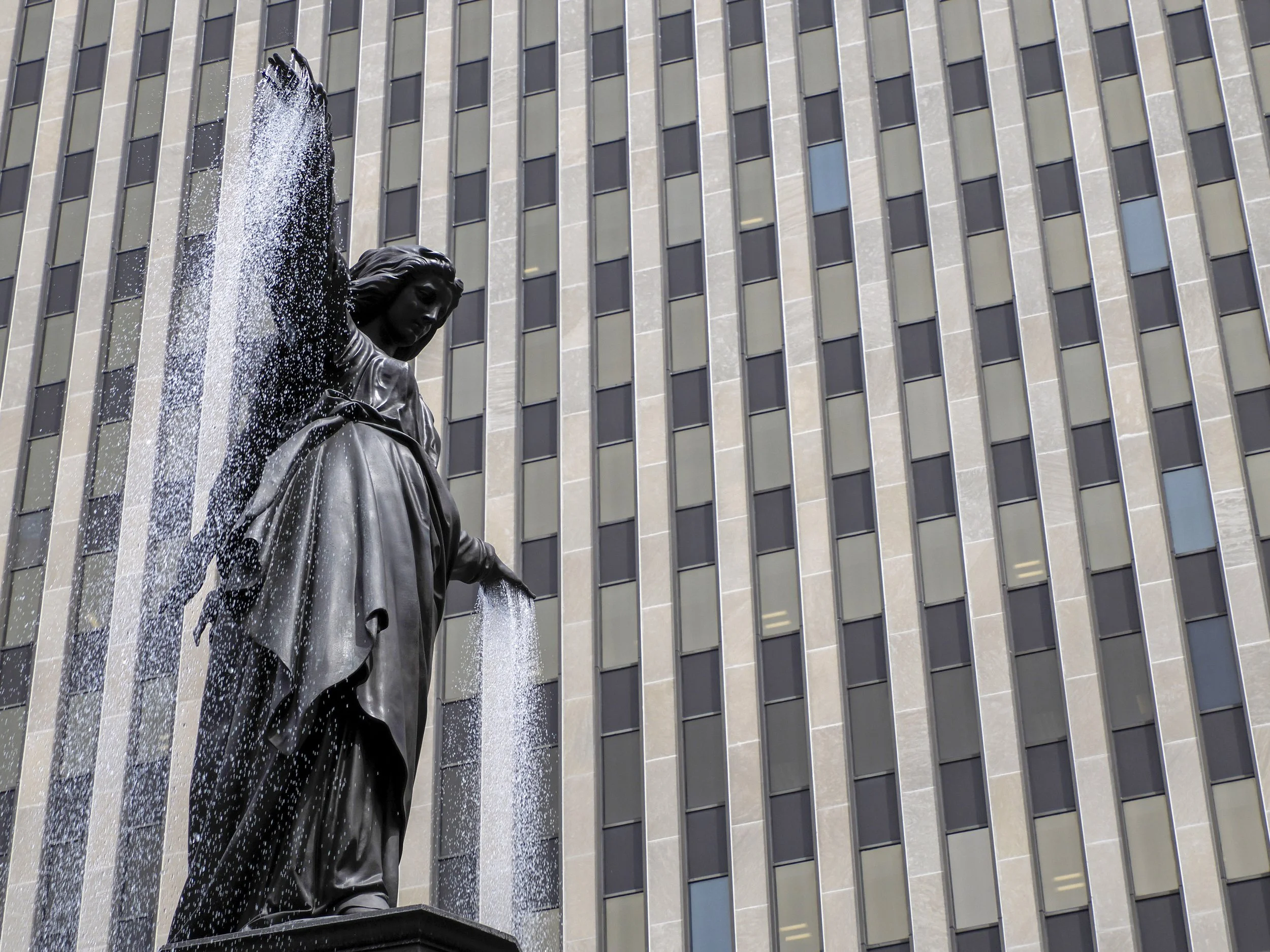 Fountain Square in Cincinnati, Ohio: a woman with water flowing from her raised arm. The background features a modern, high-rise building with a grid of windows.