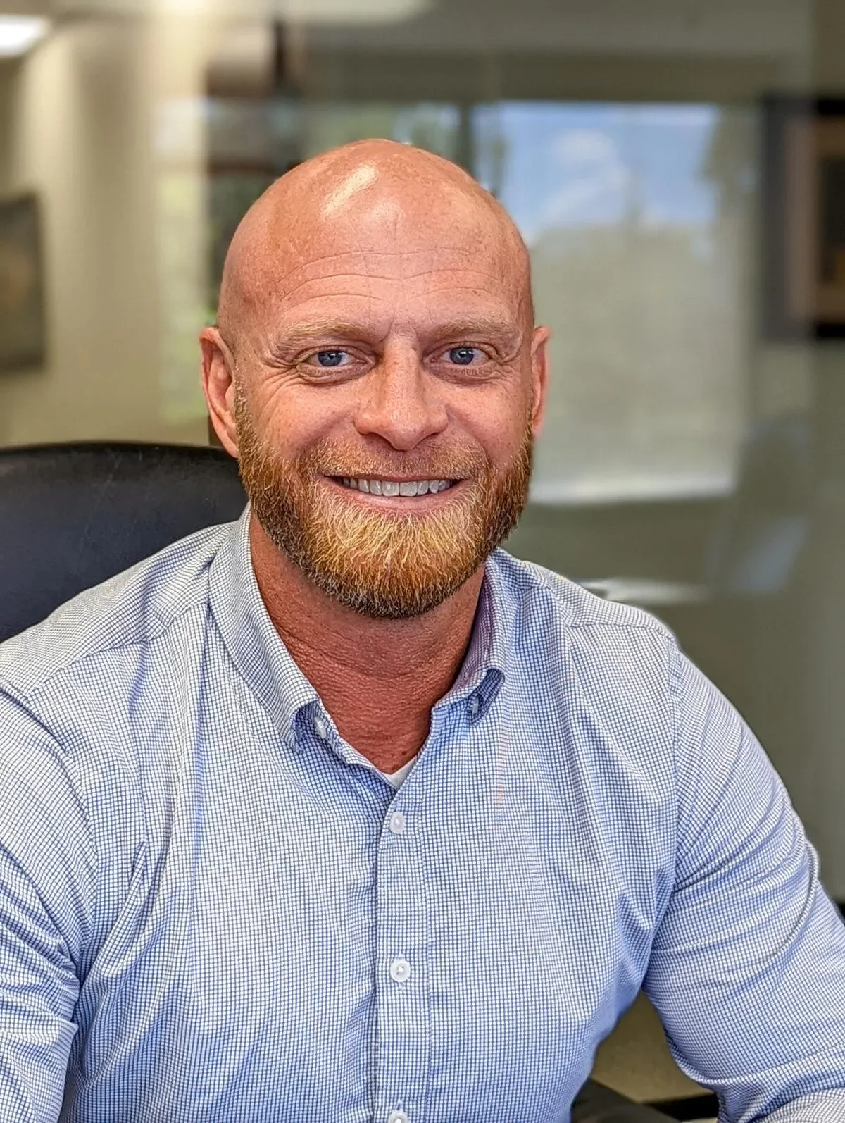 A middle-aged man with a bald head, a red beard, and blue eyes is smiling and sitting at a desk in an office. He is wearing a light blue, checkered button-down shirt.